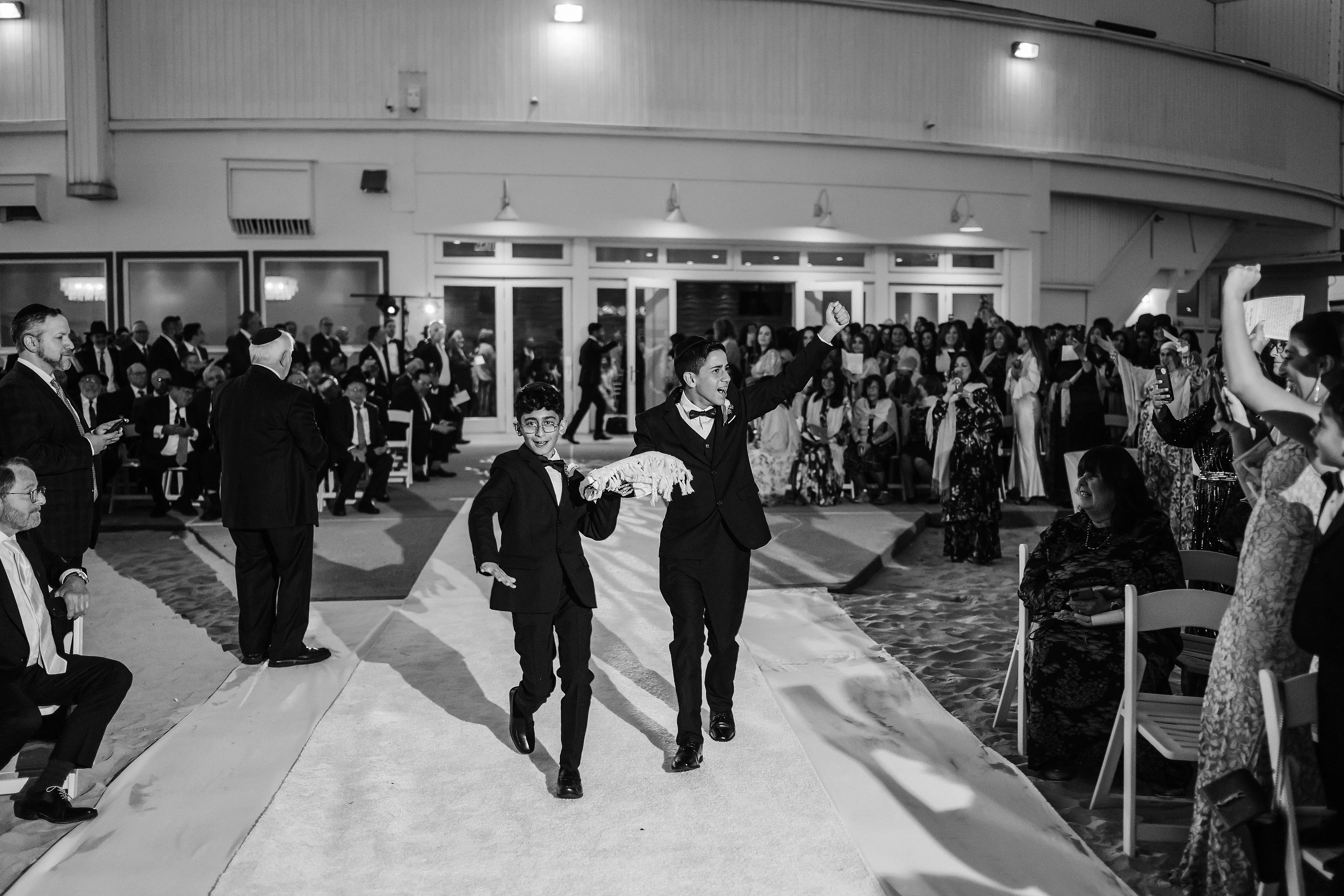 Black-and-white photo of two boys in formal attire walking forward on an aisle while guests line the sides and cheer, with raised hands and phones in the background.