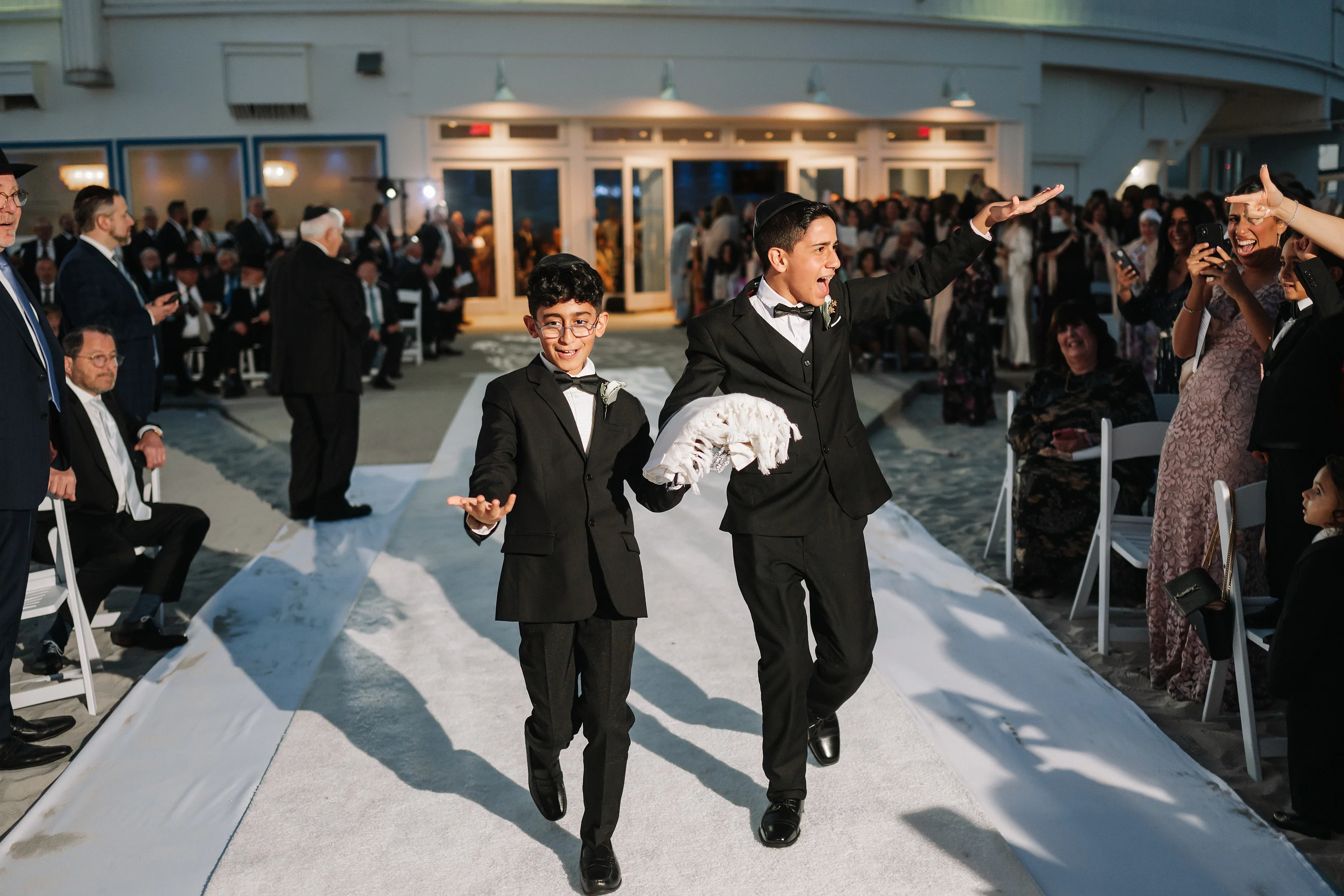 Two young celebrants in formal outfits running joyfully down the center aisle as guests applaud and watch from both sides.