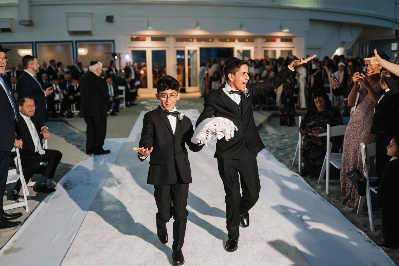 Two young celebrants in formal outfits running joyfully down the center aisle as guests applaud and watch from both sides. Wedding photography at Sands, Atlantic Beach, NY.