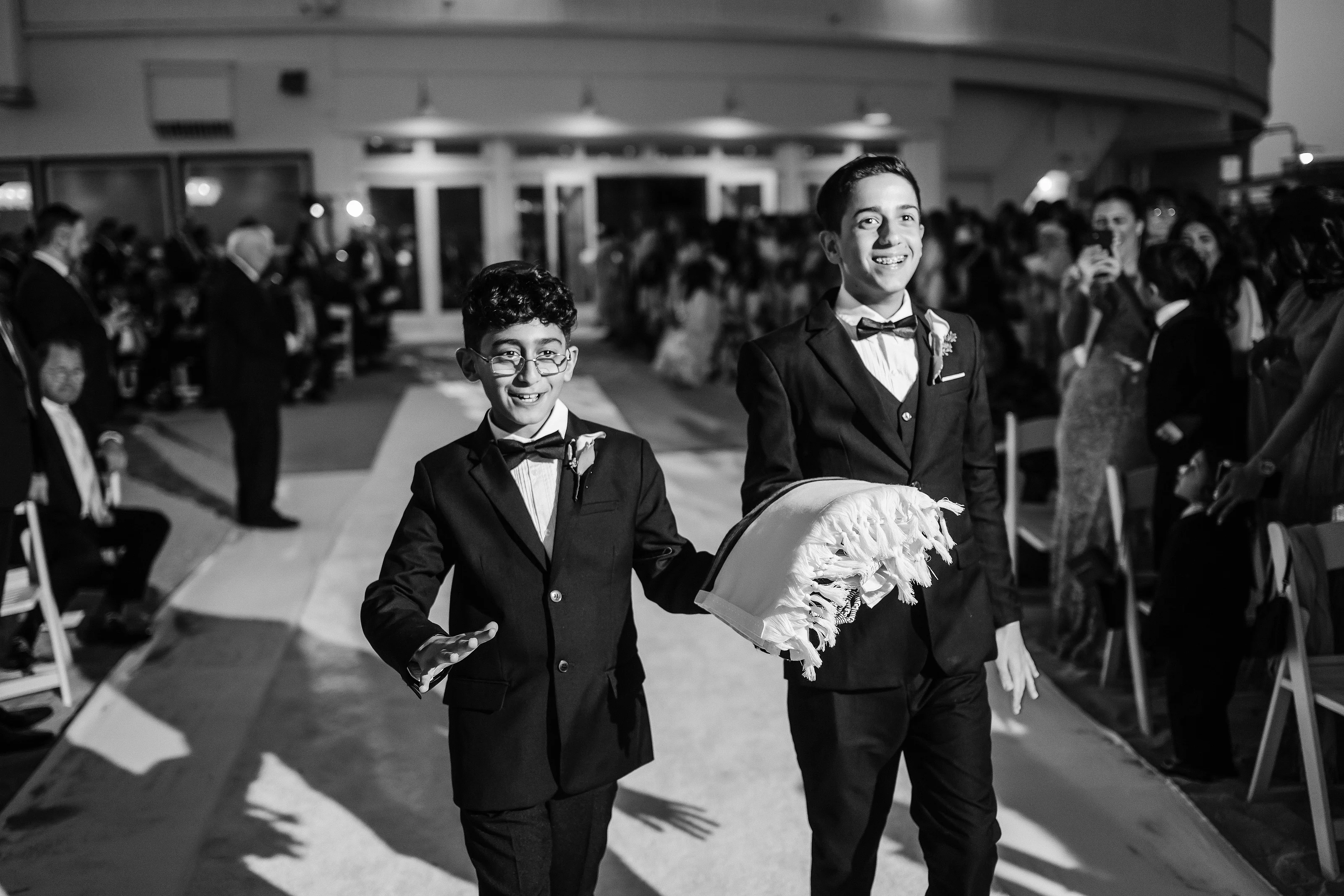 Black-and-white photo of two boys in tuxedos smiling during a lively reception, with guests seated and standing in the background.