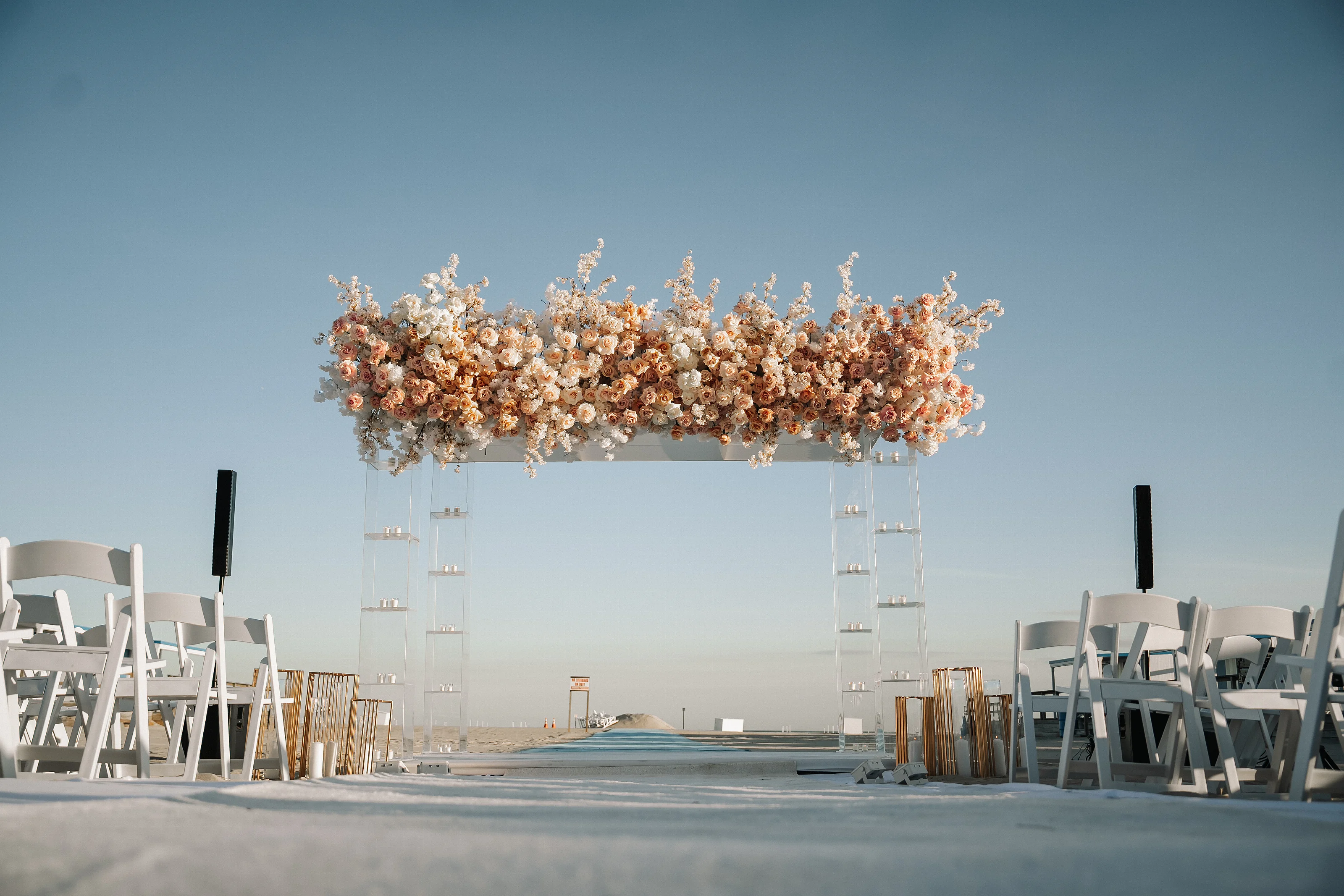 Floral chuppah canopy with hanging arrangements above a bright outdoor ceremony aisle, framed by white chairs and clear acrylic candle holders.