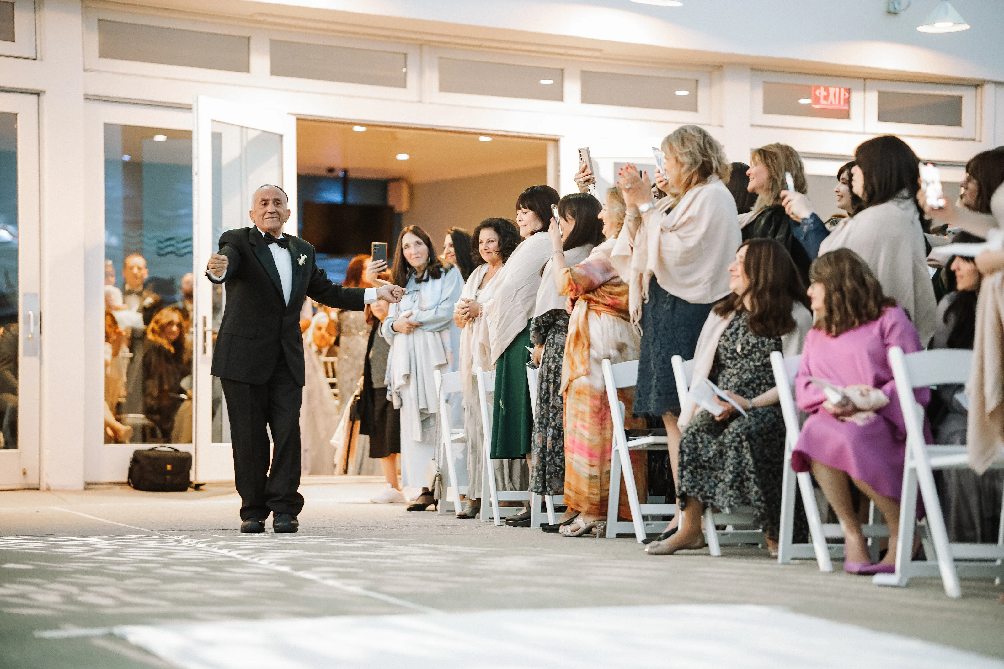 A man in a tuxedo walking down an aisle as guests seated along the sides clap and smile during a Jewish wedding celebration.