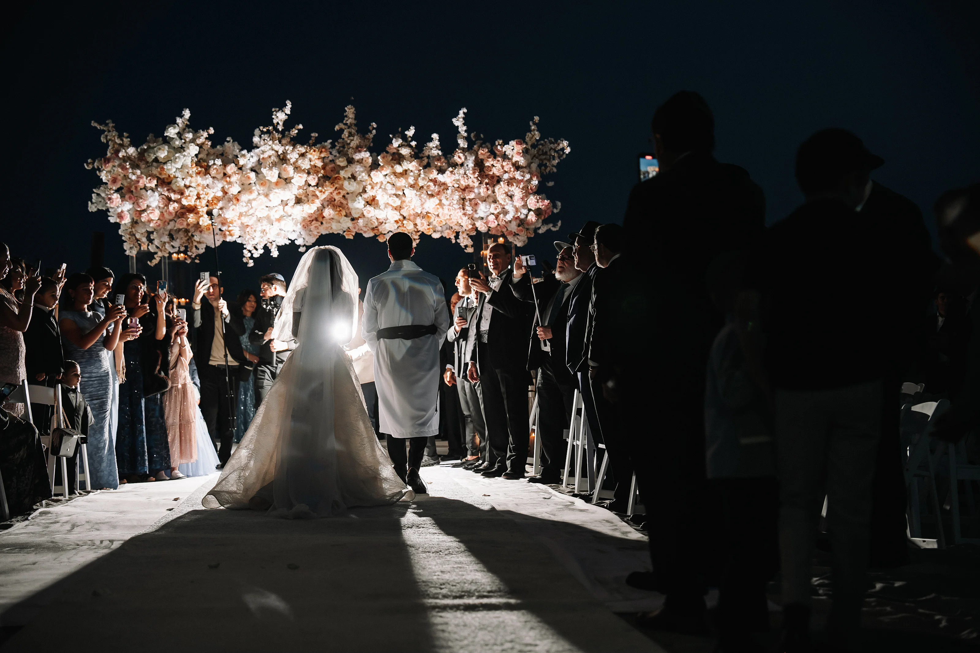 Bride and groom walking under a floral chuppah with guests lining the aisle, spotlighted against a dark night backdrop.