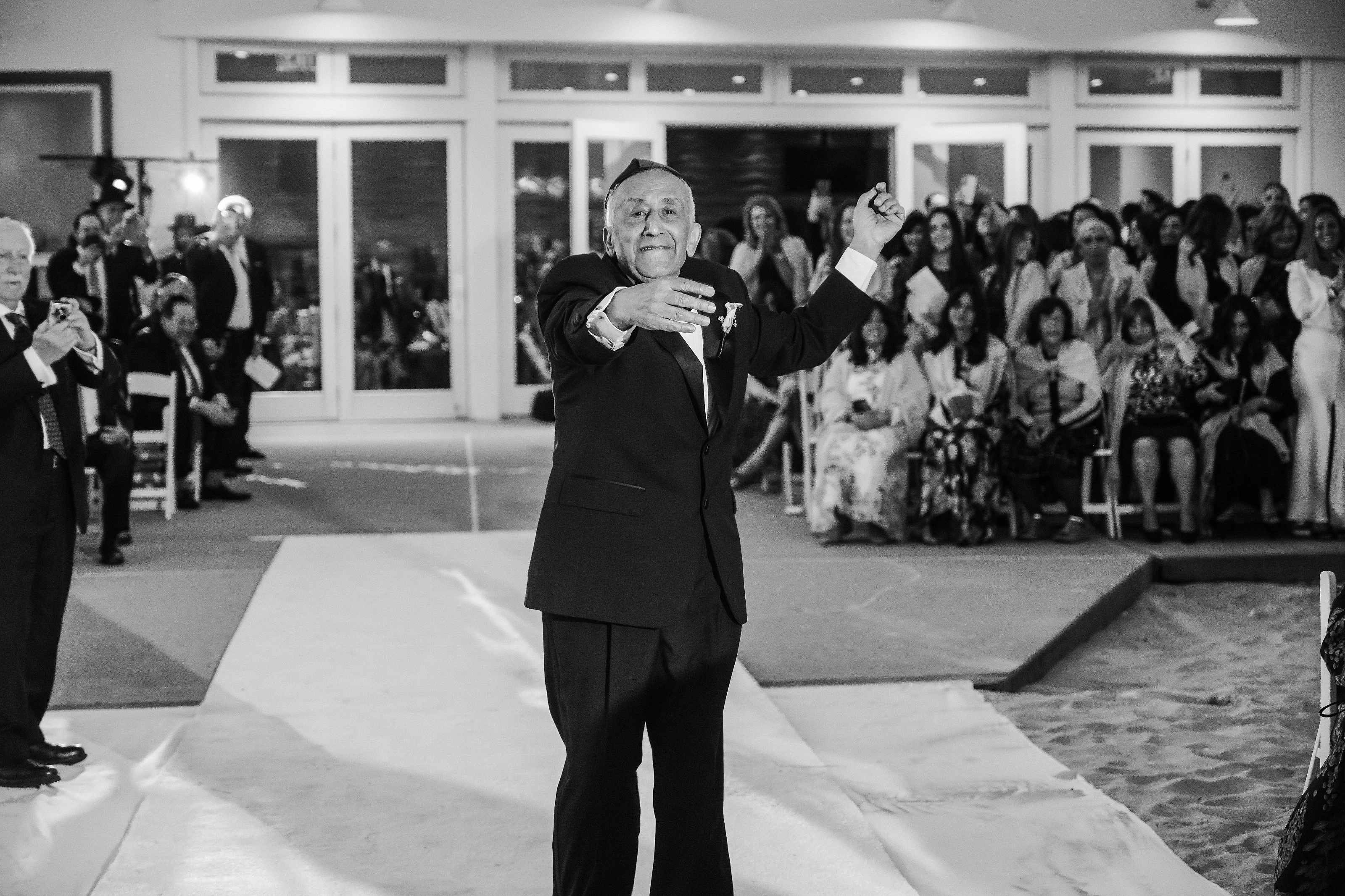 Black-and-white photo of an older man in formal attire celebrating on a wedding dance floor with arms open while guests watch from the background.