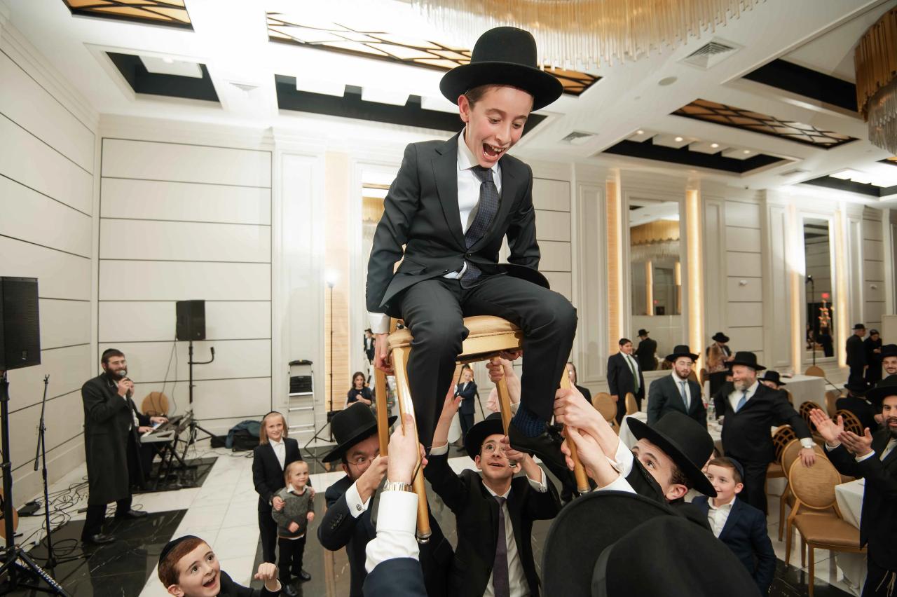 Chassidic groom-style celebrant laughing while being lifted on shoulders by guests during a wedding celebration, with other men and children cheering in the background.