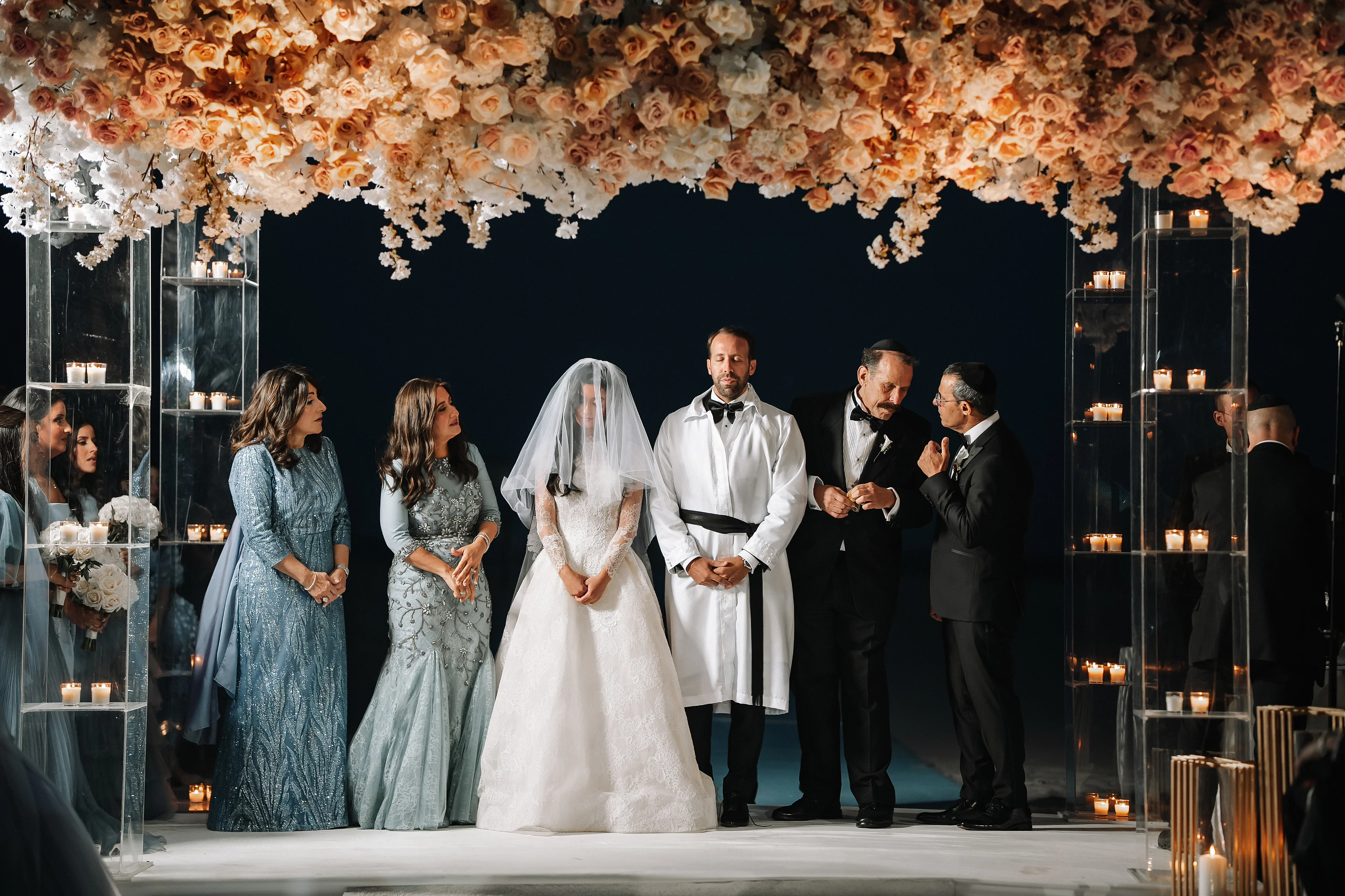 Bride in a white wedding dress under a floral chuppah canopy while family members stand nearby during the ceremony, with warm candles in glass towers framing the scene.