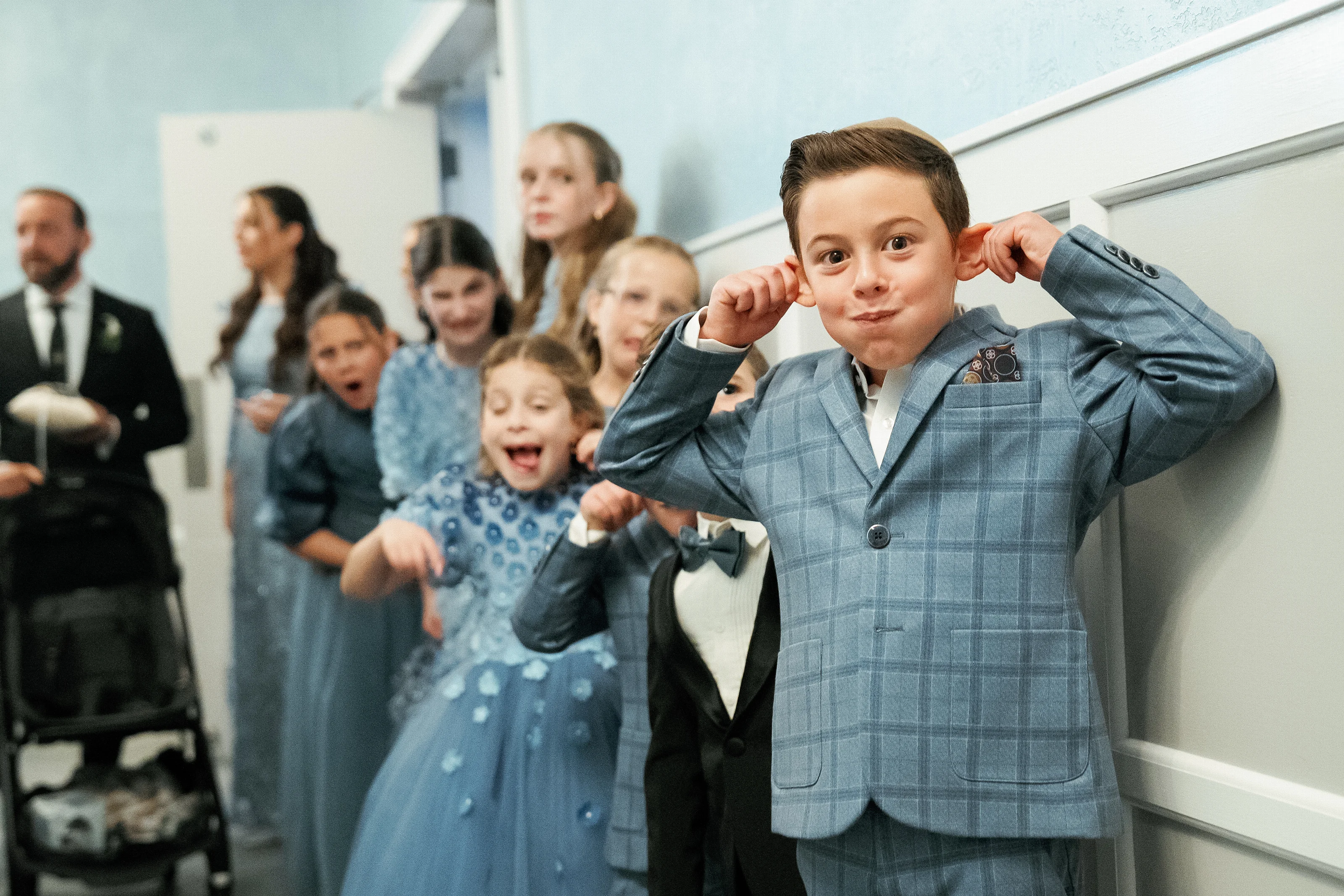 Ring bearer in a blue suit posing playfully indoors while family and guests smile in the background.