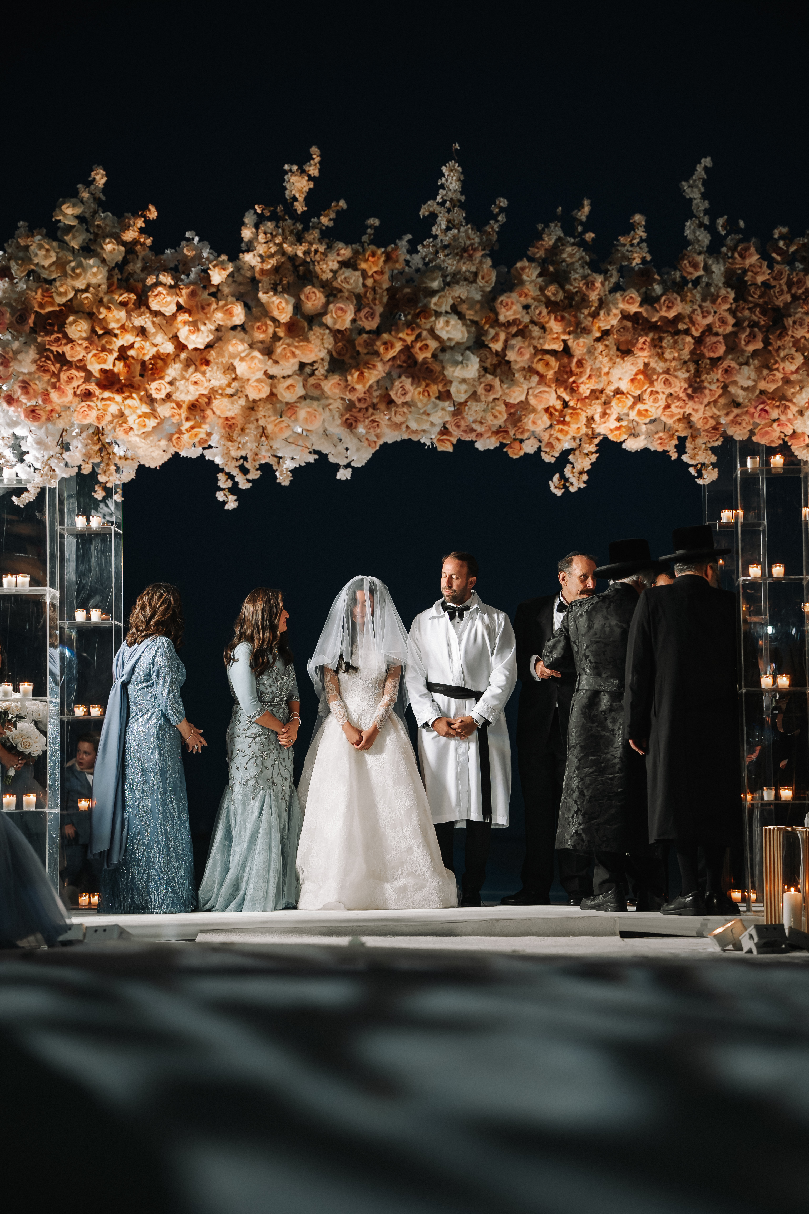 Bride and groom standing under a floral chuppah canopy at night with candlelight towers and family nearby.