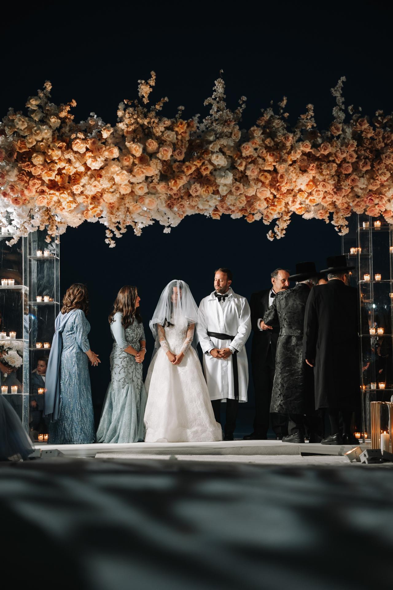 Bride and groom standing under a floral chuppah canopy at night with candlelight towers and family nearby. Wedding photography at Sands, Atlantic Beach, NY.