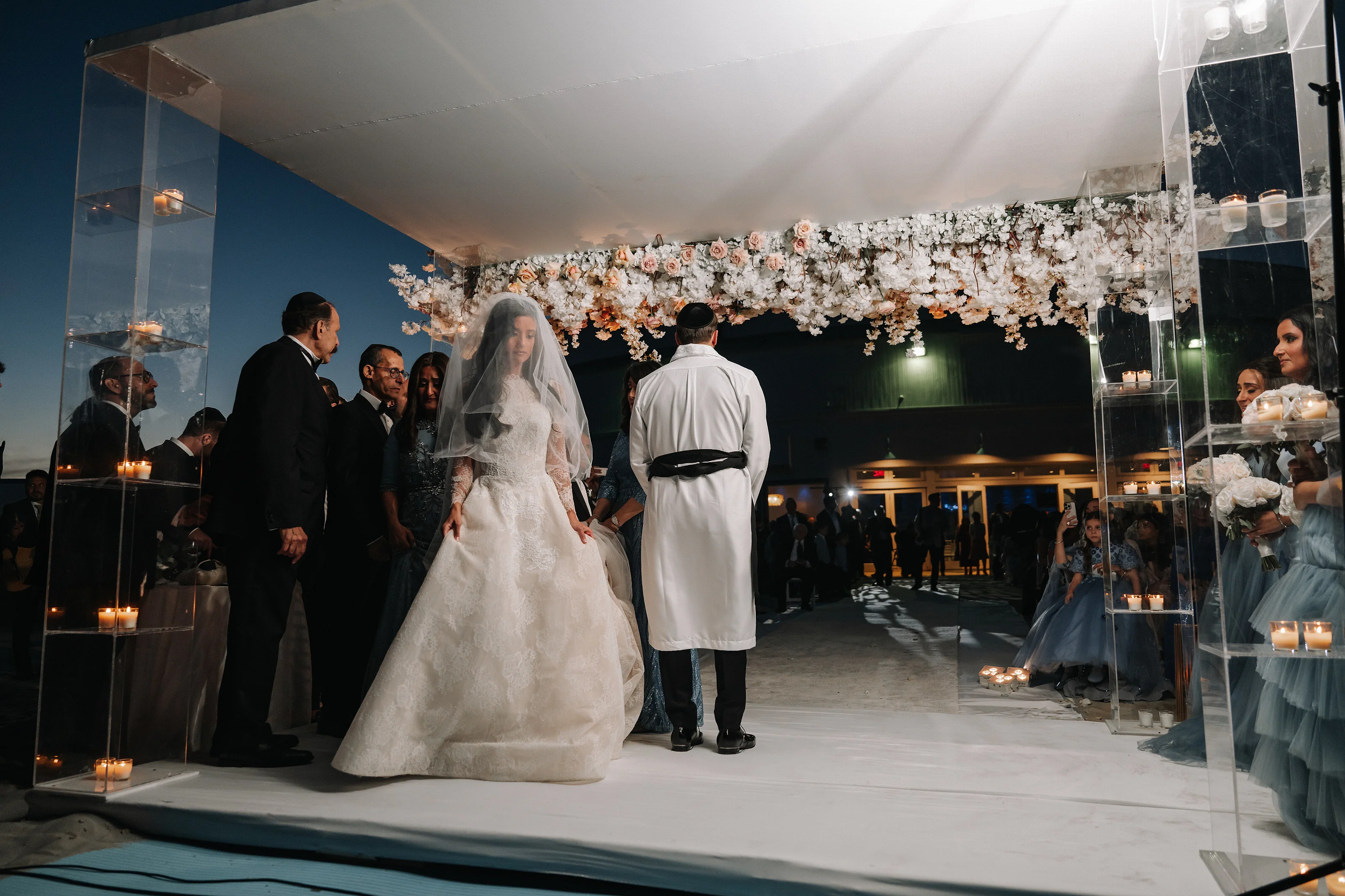 Bride in a white wedding gown and veil walking forward under a floral chuppah canopy, guests gathered on either side at a nighttime indoor wedding.