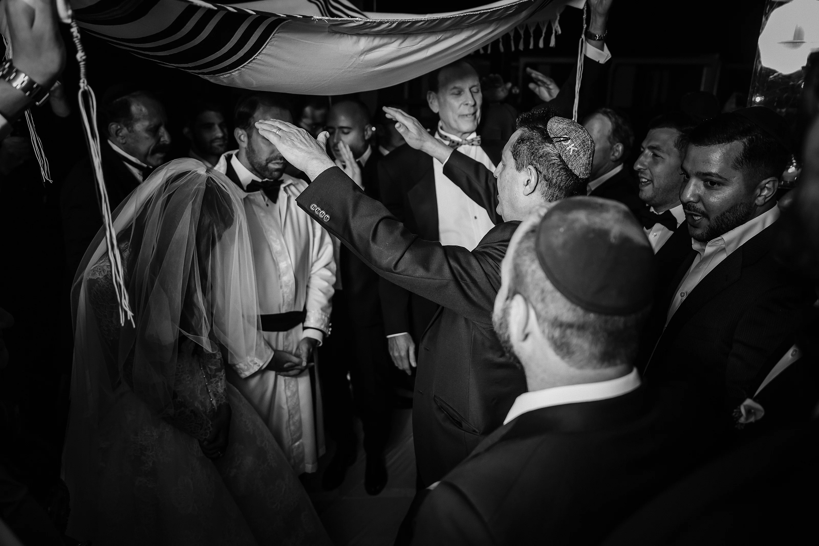 Black-and-white photo of the groom reaching toward the bride’s veil during the badeken moment with a tightly packed crowd watching.
