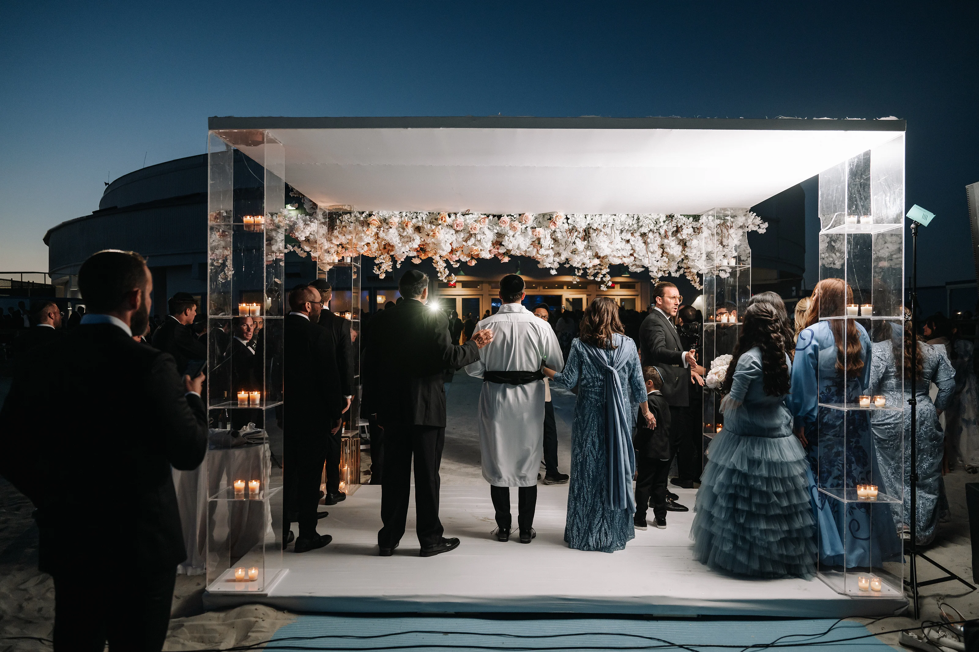 Wide night view of a chuppah canopy with floral garlands and candle-lit glass panels as guests gather on the central walkway.