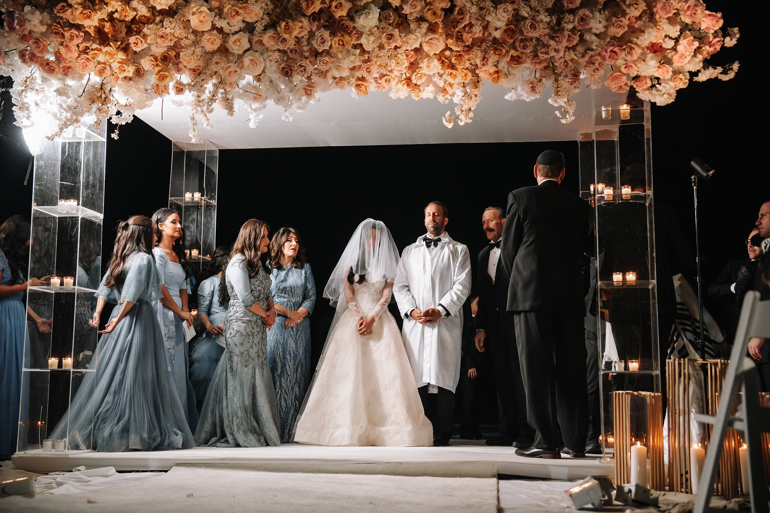 Bride and groom proceeding under a floral wedding canopy at a candlelit ceremony with family and attendants standing nearby