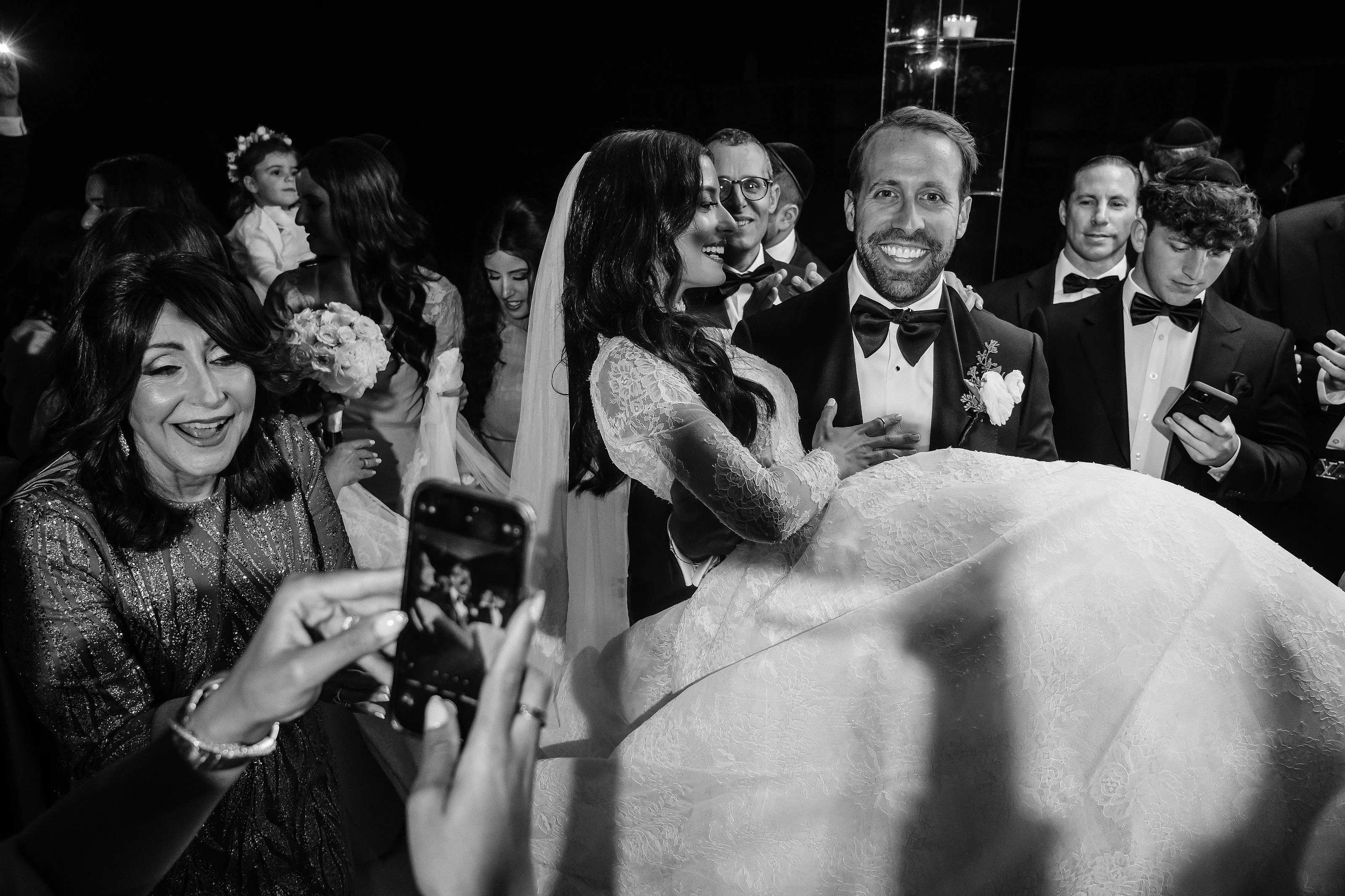 Black-and-white wedding photo of the bride and groom smiling closely as guests cheer and film with smartphones at night.