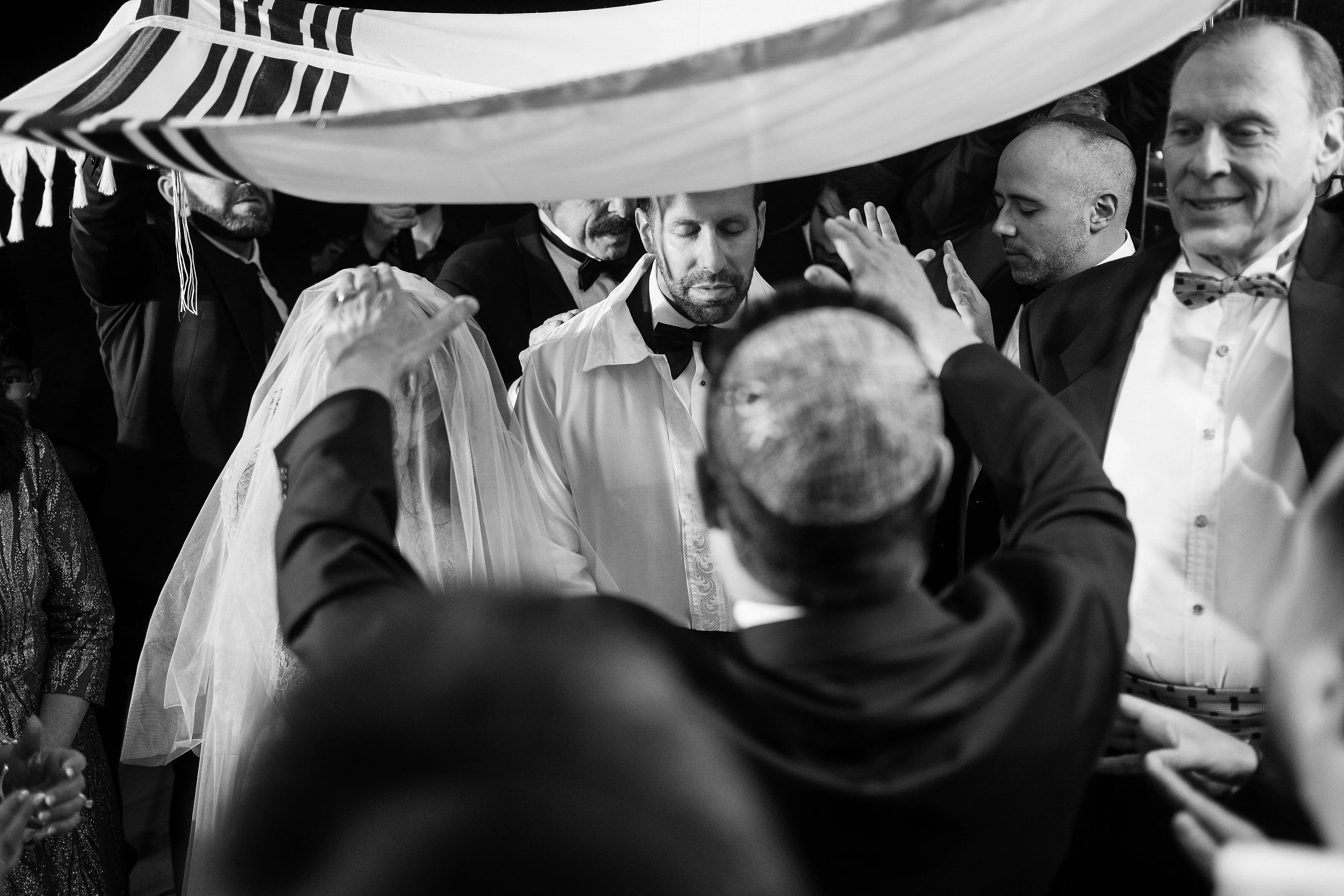 Black-and-white candid moment of a couple embracing amid a celebratory crowd under a wedding canopy, with raised hands and joyful expressions.