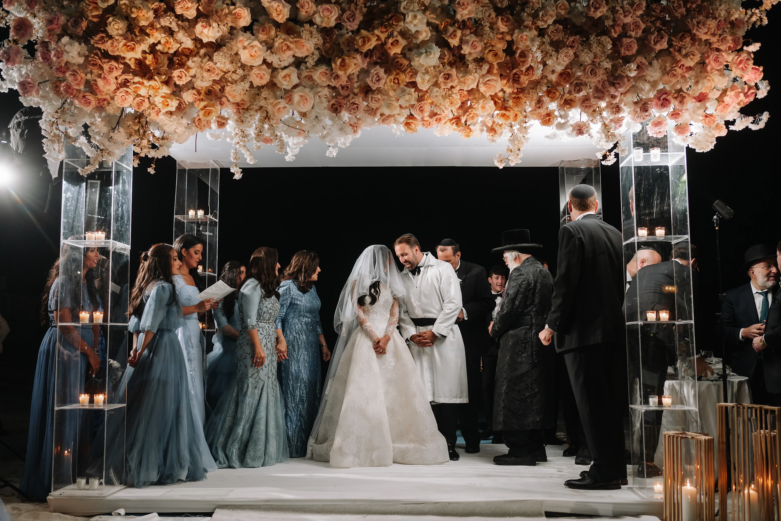 Bride and groom sharing an intimate badeken moment under a floral canopy and candlelit chuppah, with guests gathered at the sides.