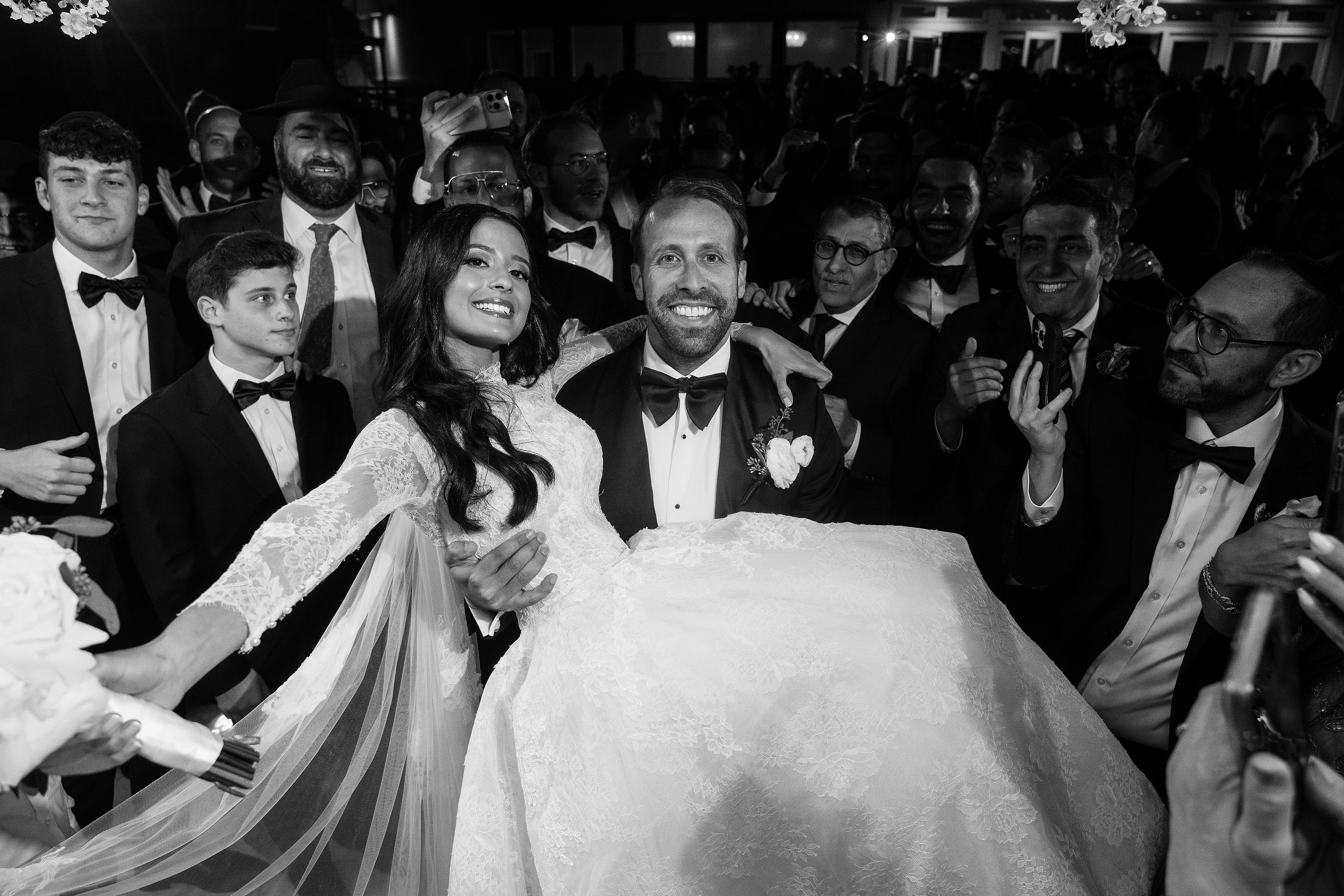 Black-and-white candid wedding moment of the bride and groom in the foreground surrounded by cheering guests during a joyful celebration.