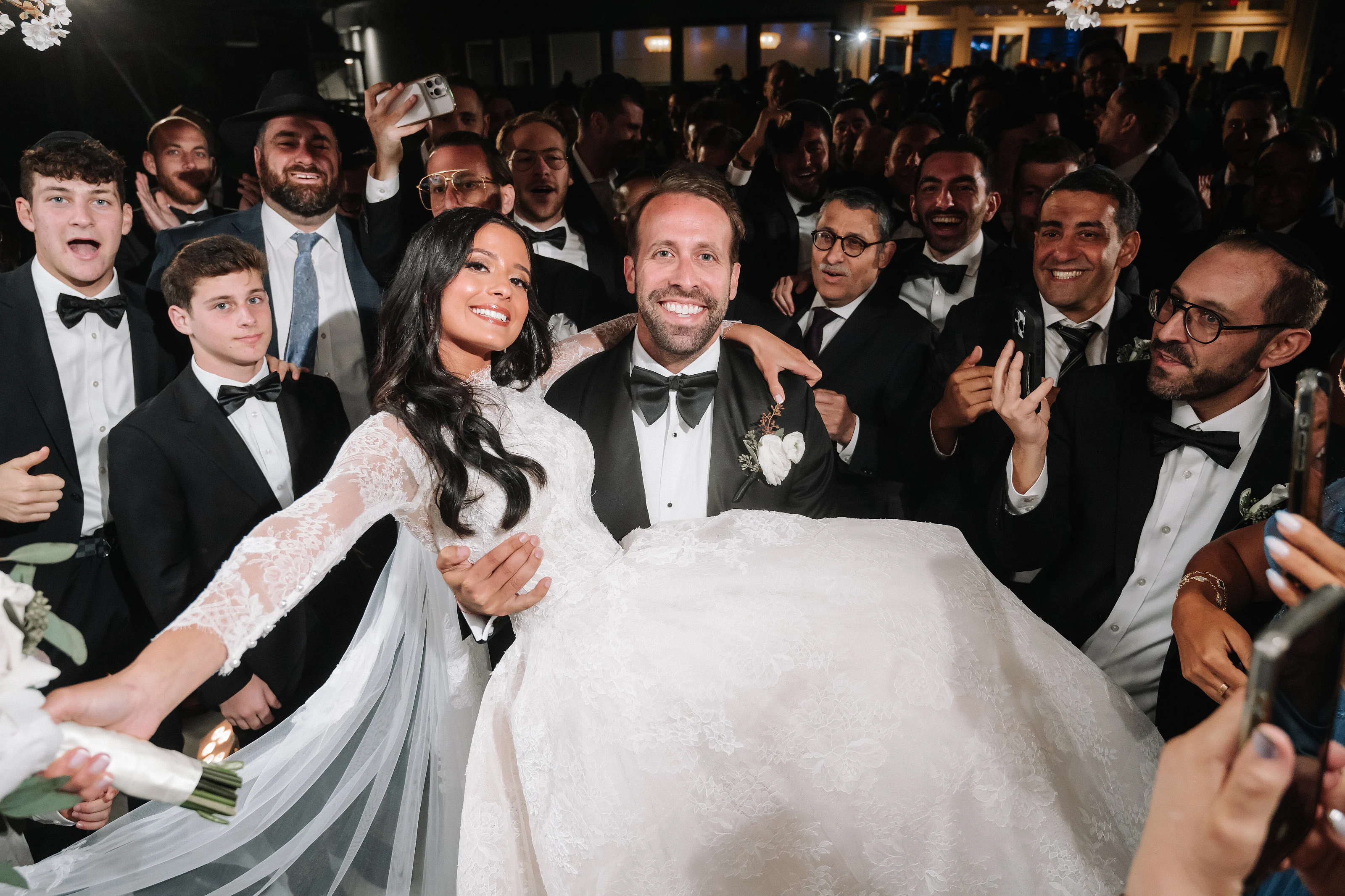 Bride in white gown being lifted by the groom during a joyful celebration while guests in tuxedos clap and cheer at an indoor event.