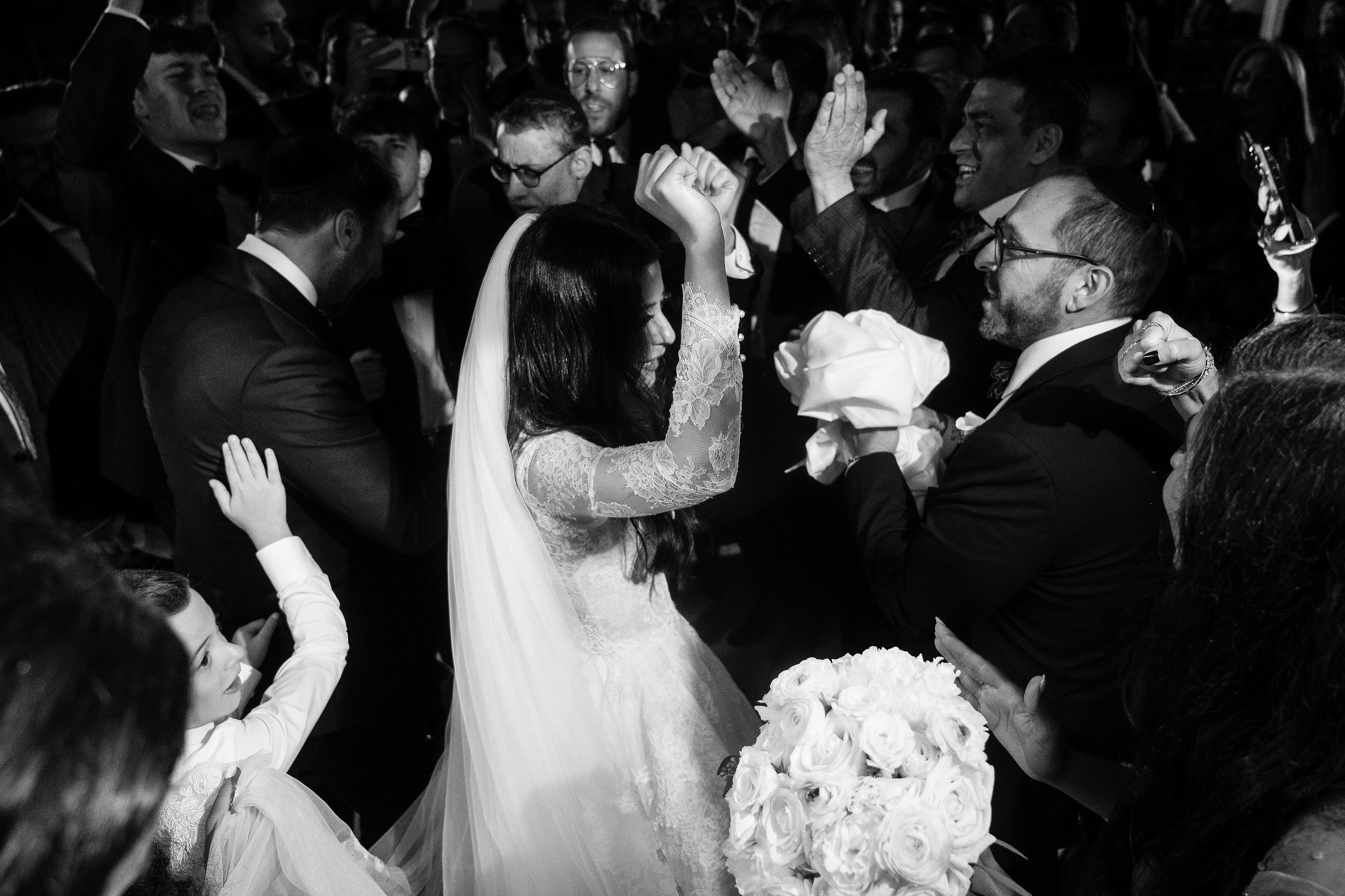 Black-and-white ceremony moment where the bride’s veil is placed as the groom leans in, surrounded by close family and guests.