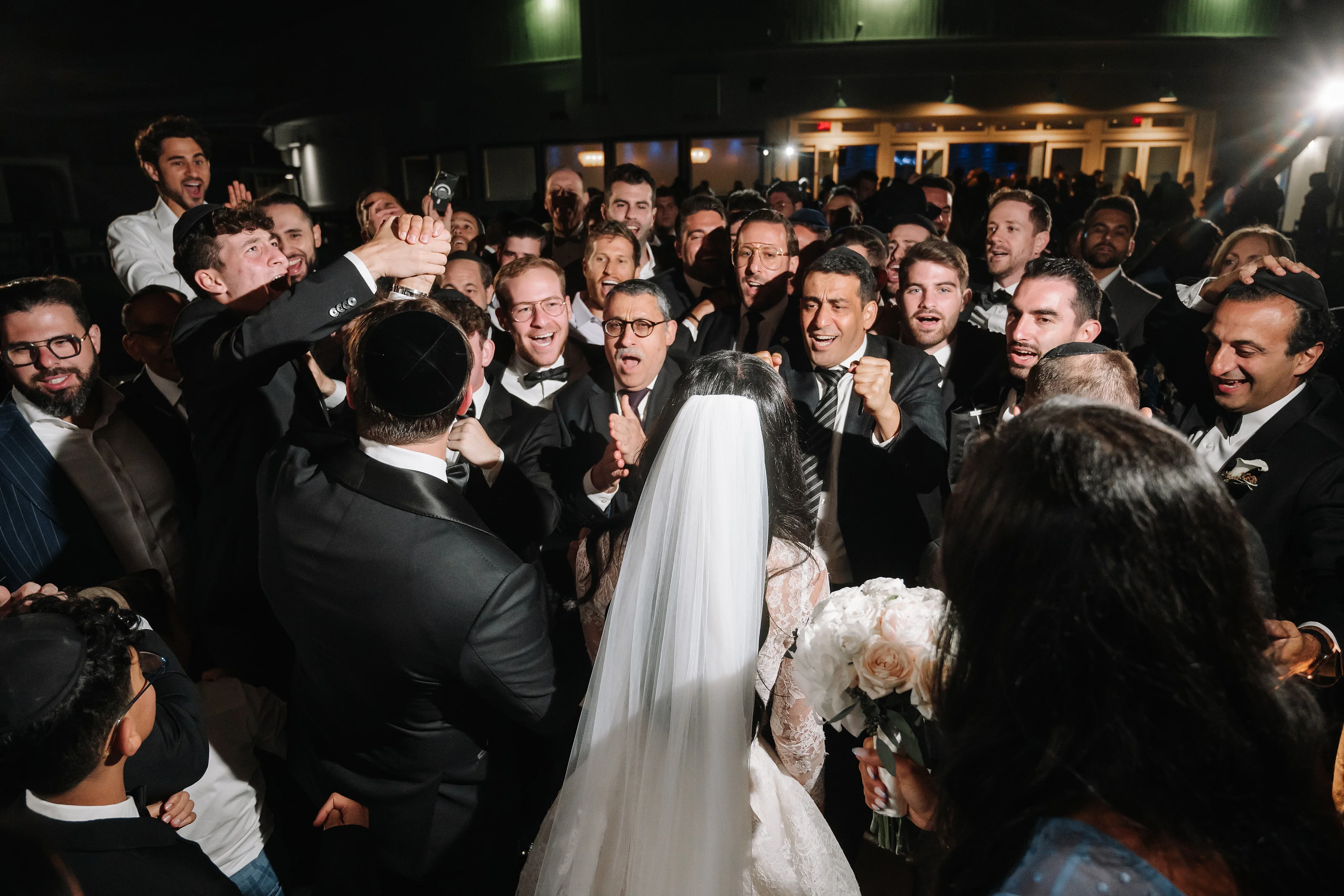 Bride in a veil holding a bouquet as the groom and a crowd of guests cheer and celebrate at a wedding reception.