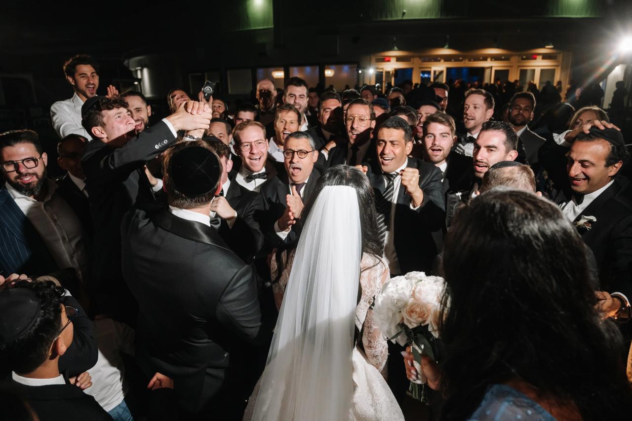 Bride in a veil holding a bouquet as the groom and a crowd of guests cheer and celebrate at a wedding reception. Wedding photography at Sands, Atlantic Beach, NY.