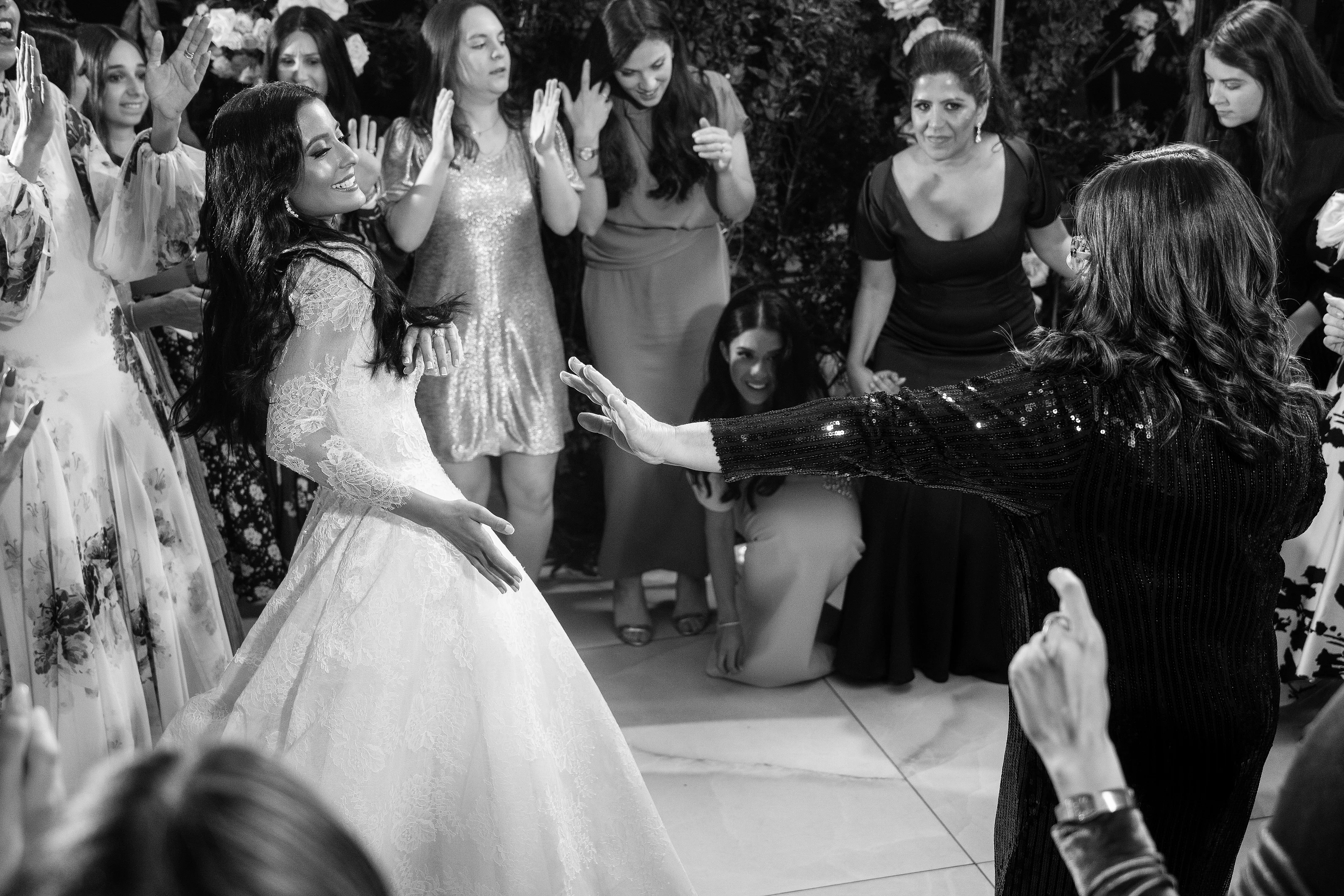 Black-and-white photo of a bride dancing with her arms extended while guests clap and watch in celebration.