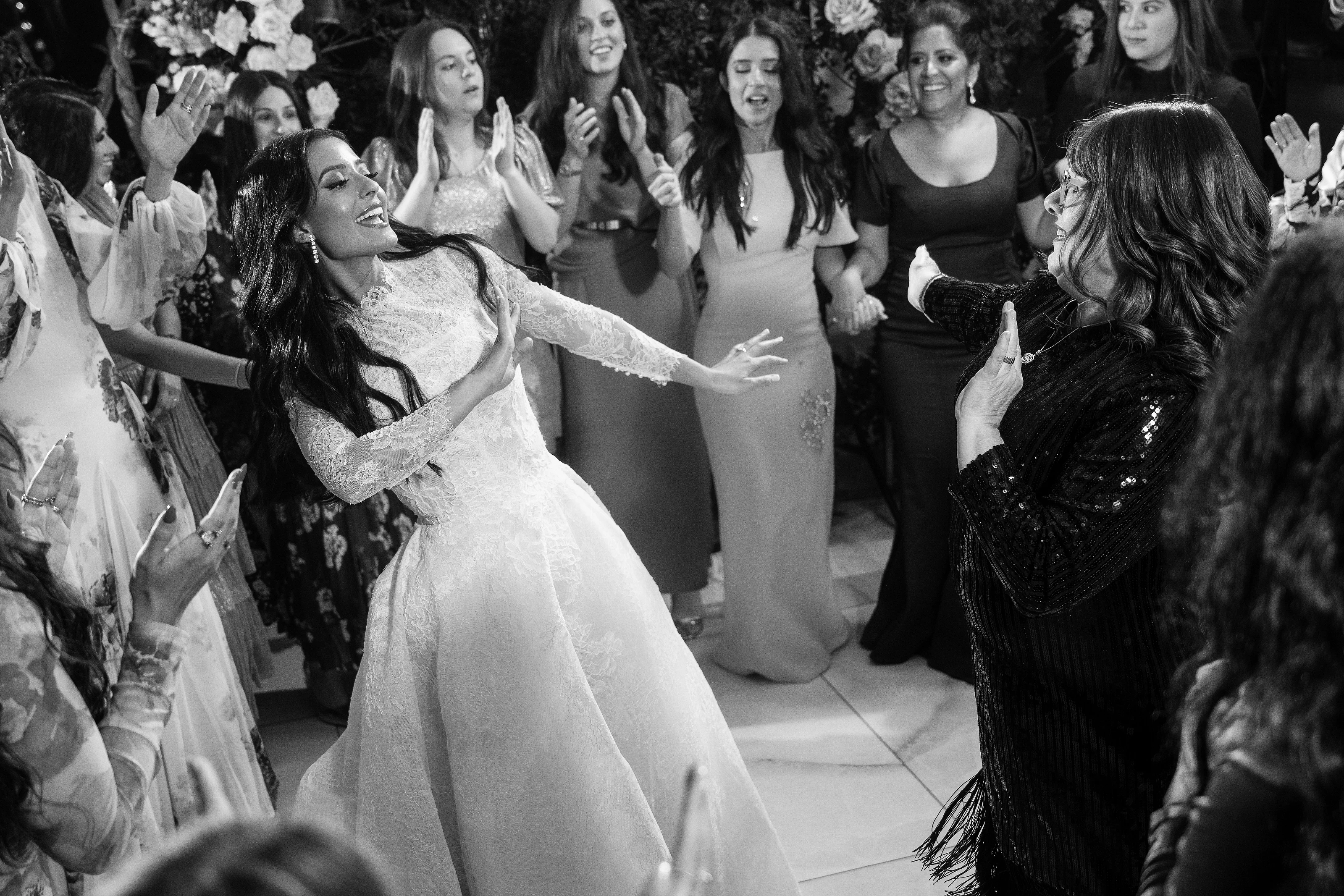 Black-and-white wedding dance-floor moment: bride smiling mid-turn while guests clap and cheer around her.