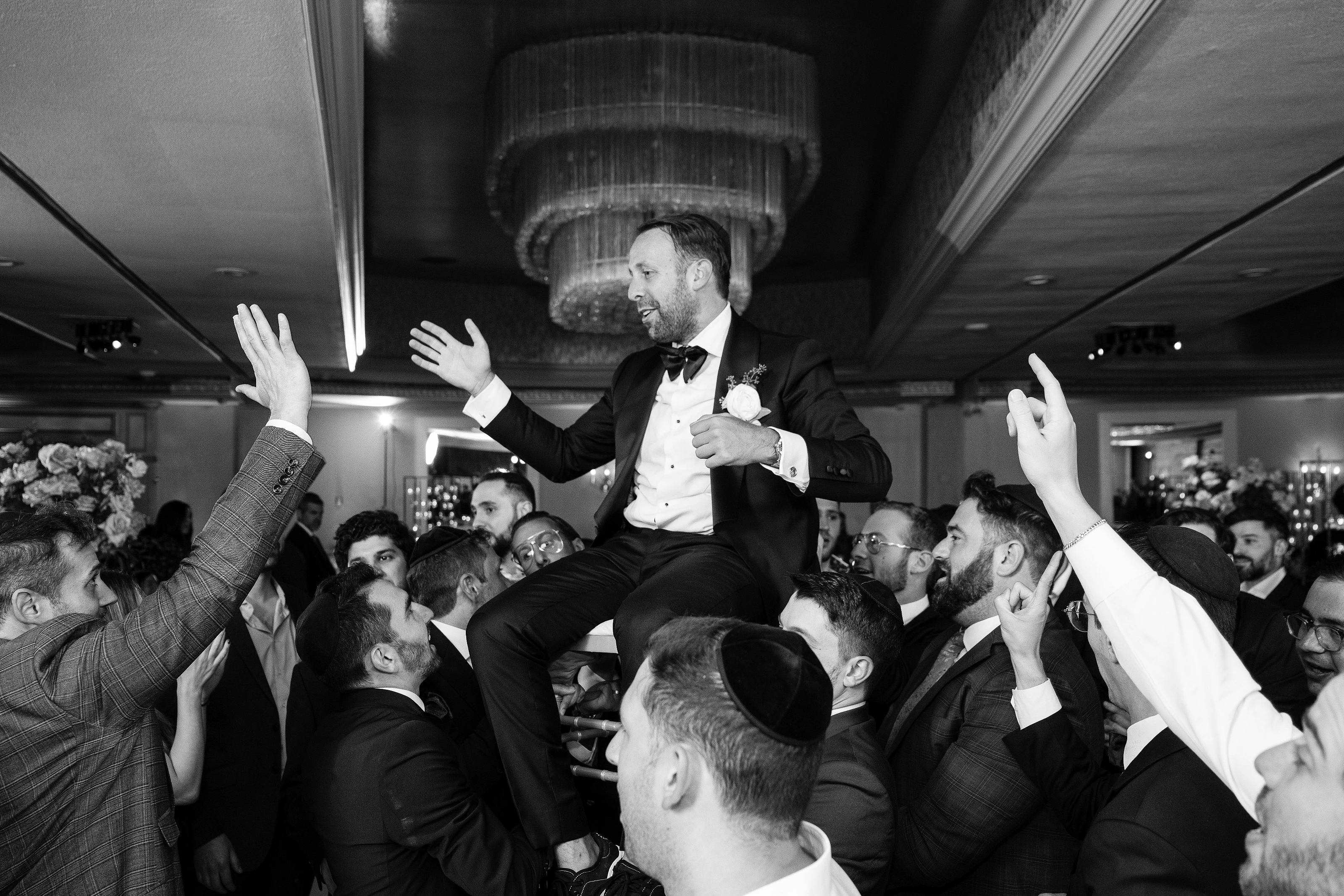 Black-and-white wedding reception photo of the groom in a tuxedo elevated above the crowd, mid-step with hands raised, surrounded by guests during dancing.
