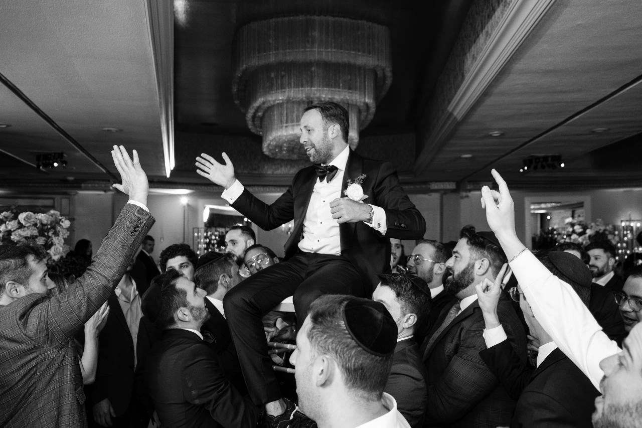 Black-and-white wedding reception photo of the groom in a tuxedo elevated above the crowd, mid-step with hands raised, surrounded by guests during dancing. Wedding photography at Sands, Atlantic Beach, NY.