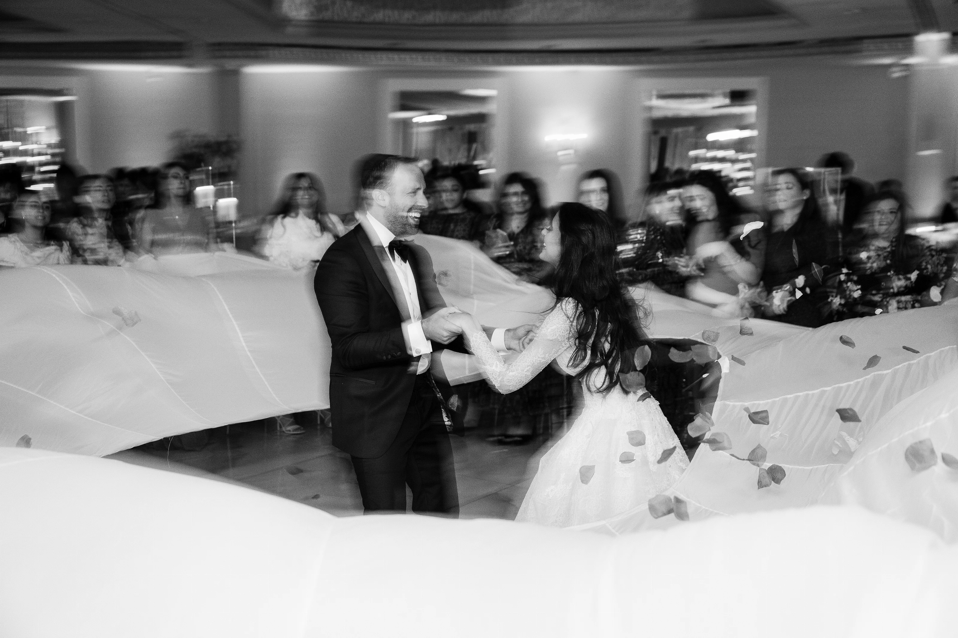 Black-and-white reception photo of a bride and groom dancing closely together with confetti while guests blur in the background.