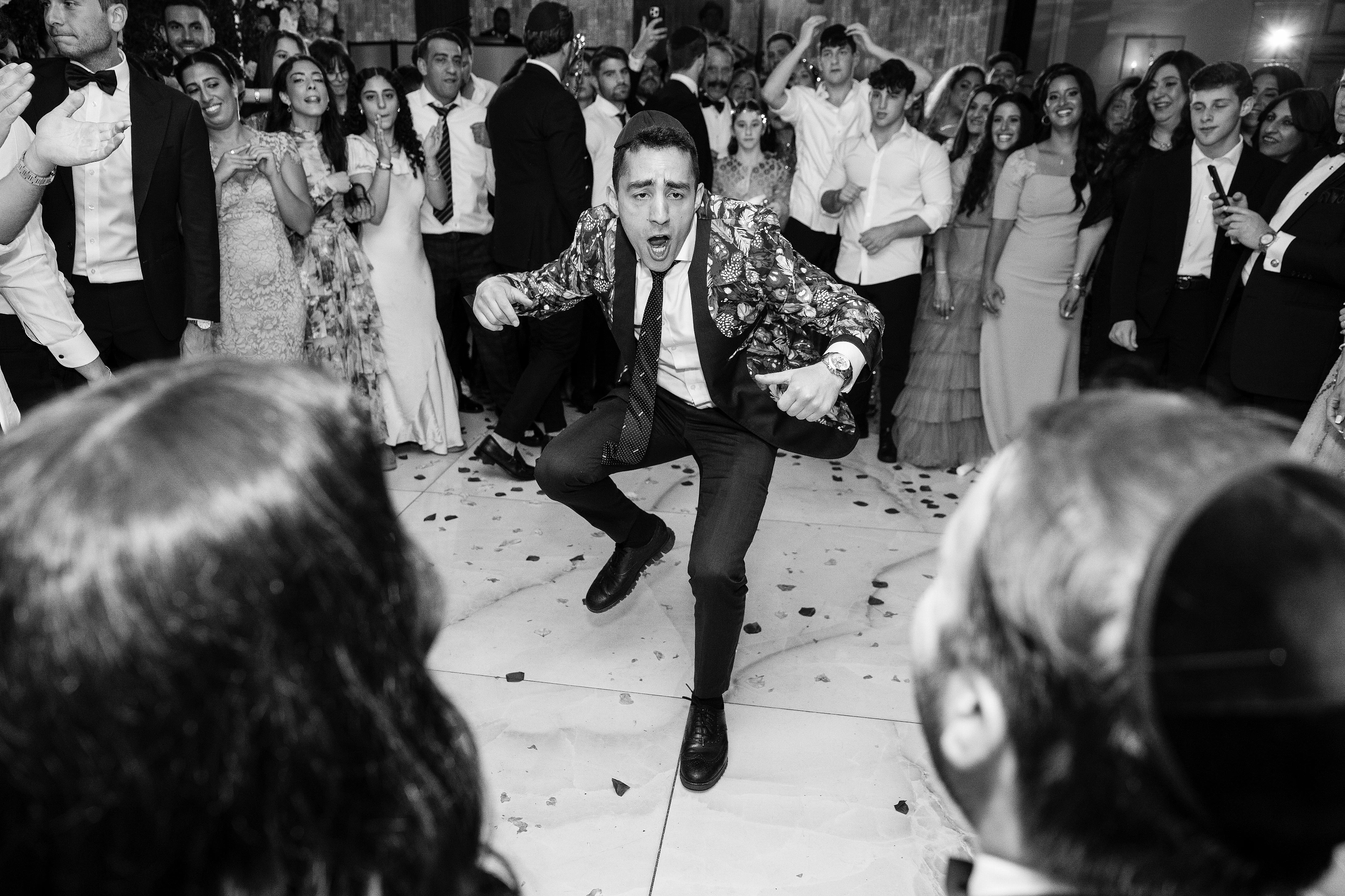 Black-and-white wedding dance-floor moment with the groom mid-run and mid-shout, confetti on the floor, and a cheering crowd in the background.