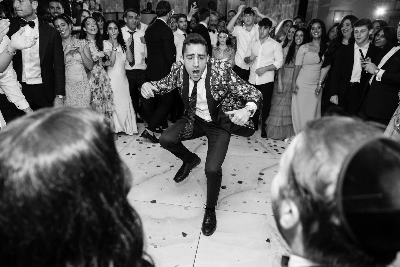 Black-and-white wedding dance-floor moment with the groom mid-run and mid-shout, confetti on the floor, and a cheering crowd in the background. Wedding photography at Sands, Atlantic Beach, NY.