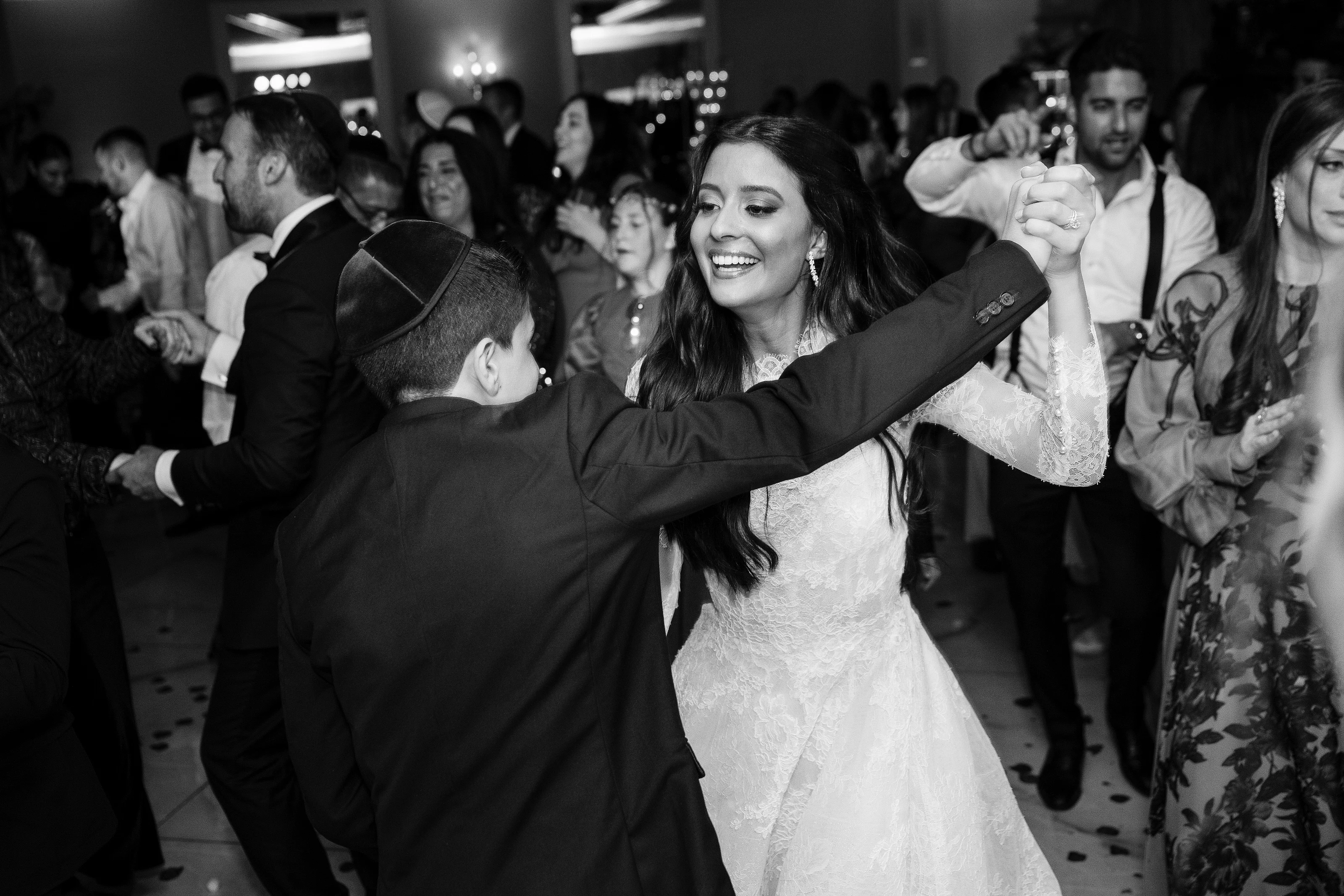 Black-and-white reception photo of a bride laughing while being turned by the groom during the celebration, with confetti on the dance floor and guests watching in the background.