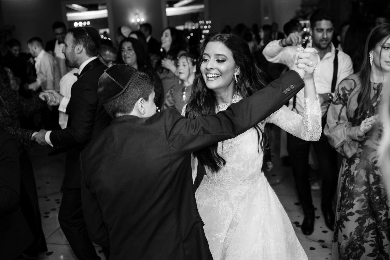Black-and-white reception photo of a bride laughing while being turned by the groom during the celebration, with confetti on the dance floor and guests watching in the background. Wedding photography at Sands, Atlantic Beach, NY.