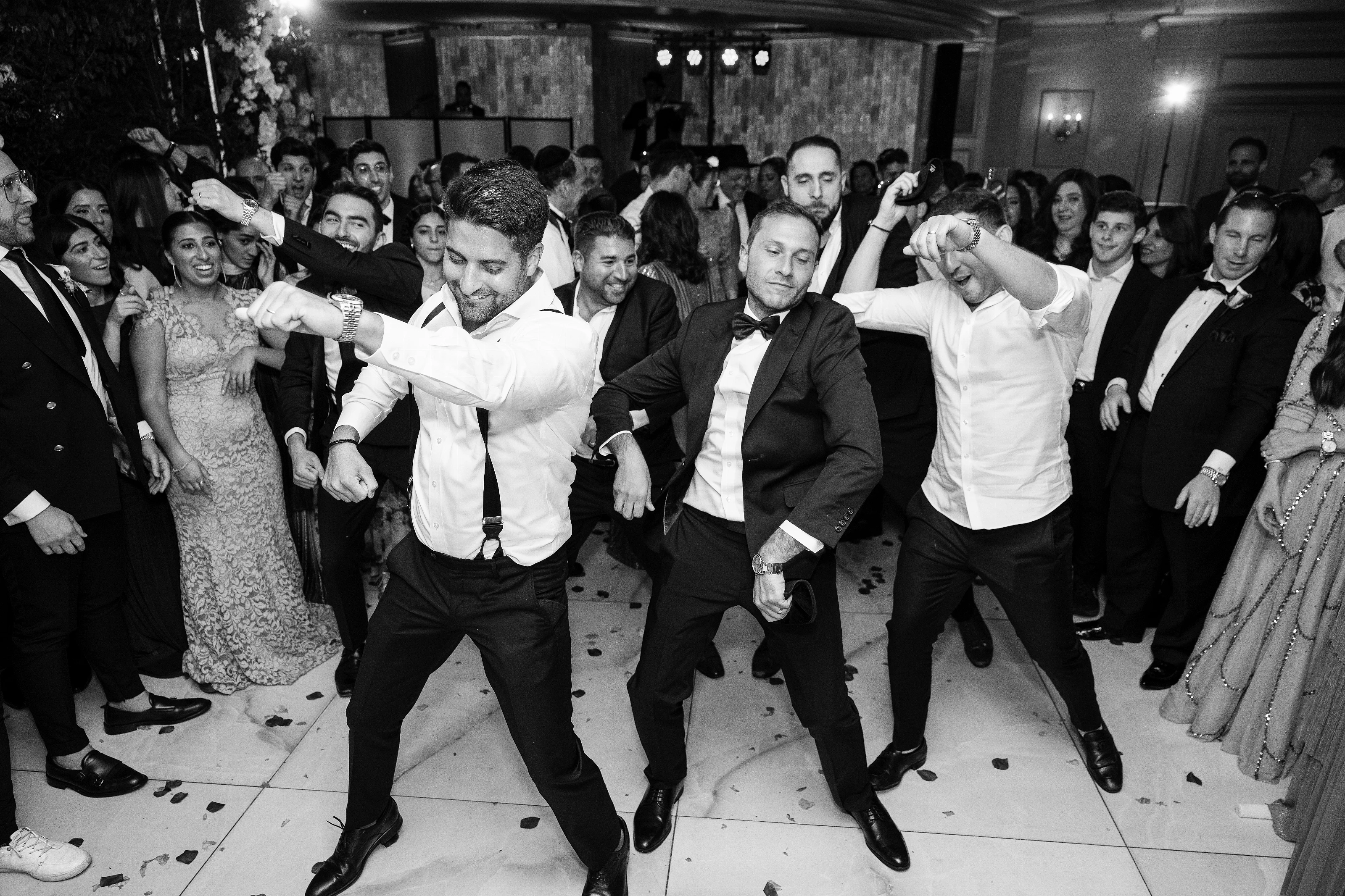 Black-and-white wedding reception photo of a groom dancing mid-move with guests cheering and confetti on the floor.