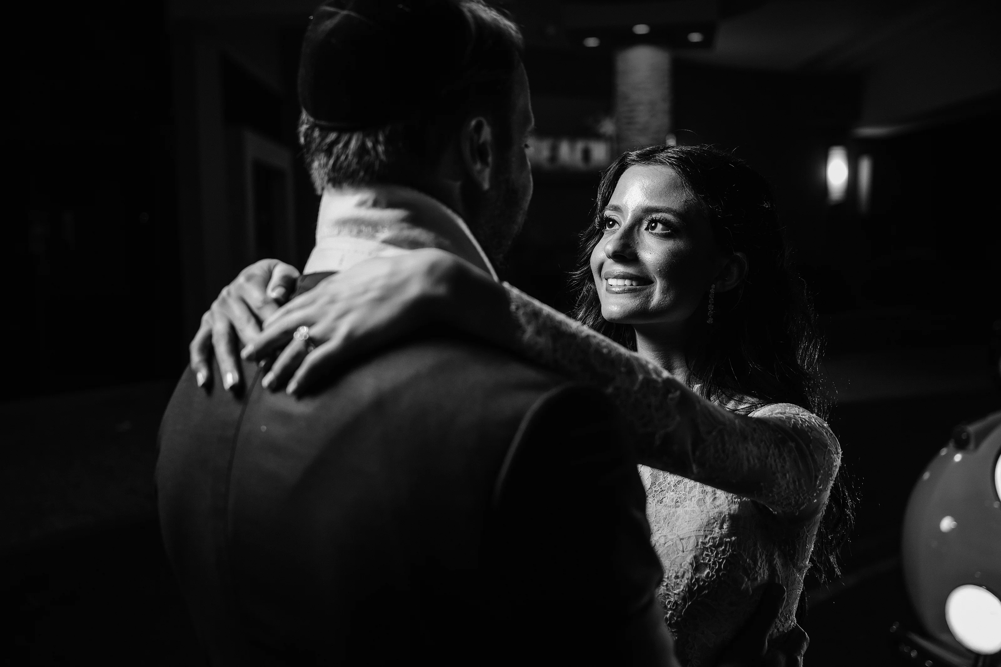 Black-and-white wedding reception moment: smiling bride embracing groom in a close hug amid dark ambient lighting and soft highlights.