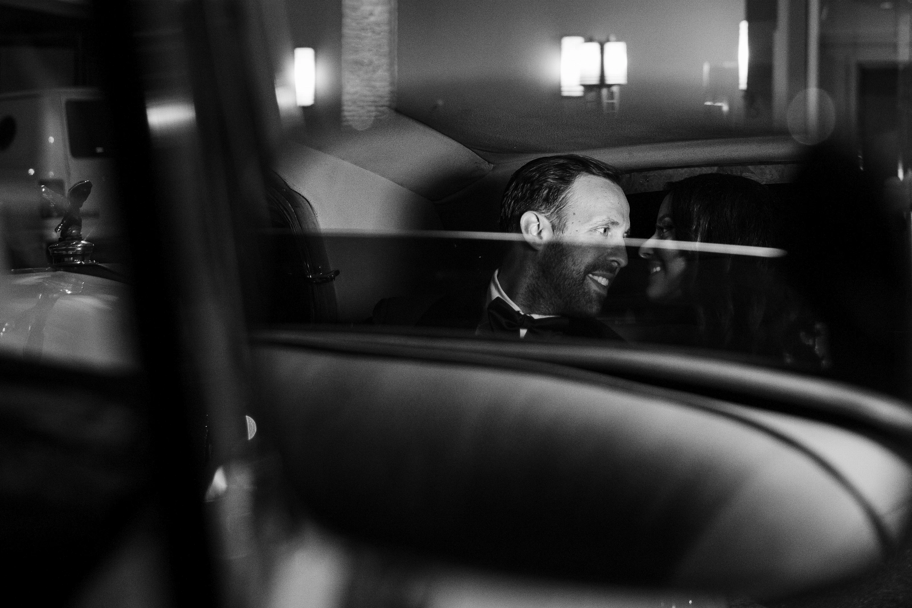 Black-and-white wedding photo of a groom looking off to the side from behind a reflective foreground, with soft out-of-focus lights in the background.