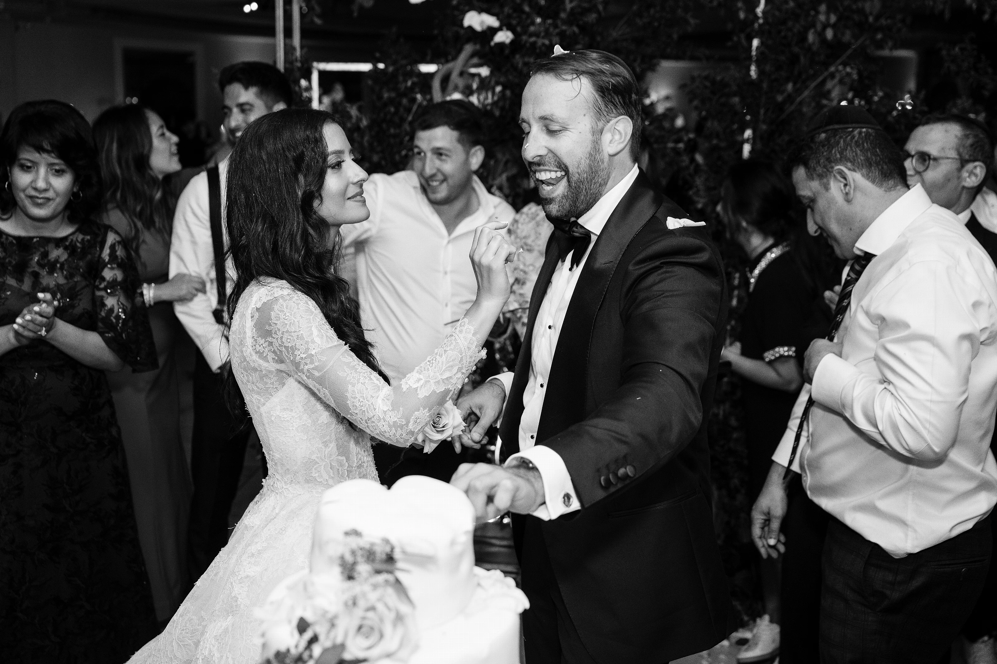 Black-and-white candid of a bride and groom smiling and holding hands during the reception while guests surround them in celebration.