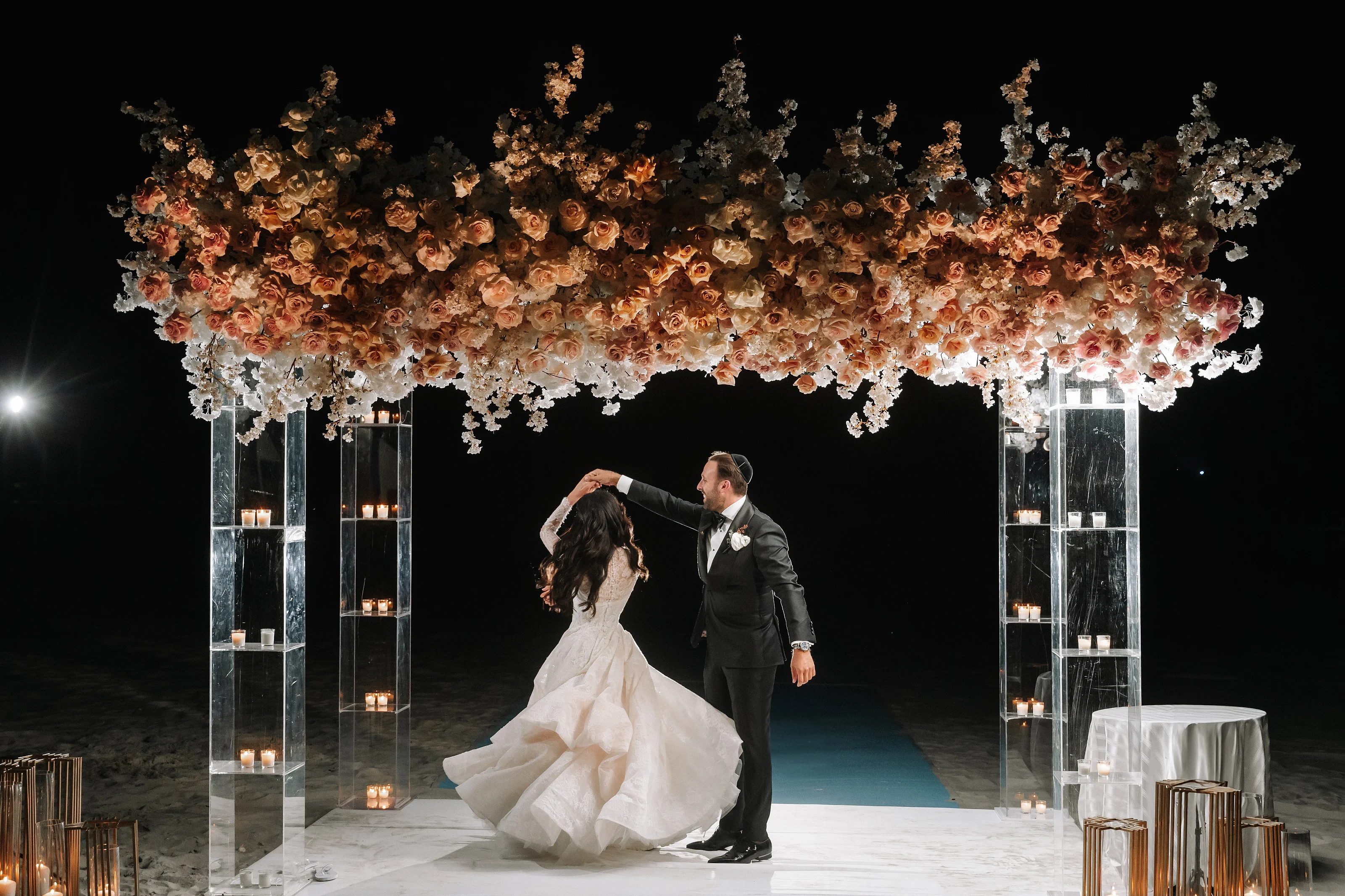 Bride and groom beneath a floral chuppah arch with warm candles and dramatic spotlights, the bride’s veil flowing as they move together.