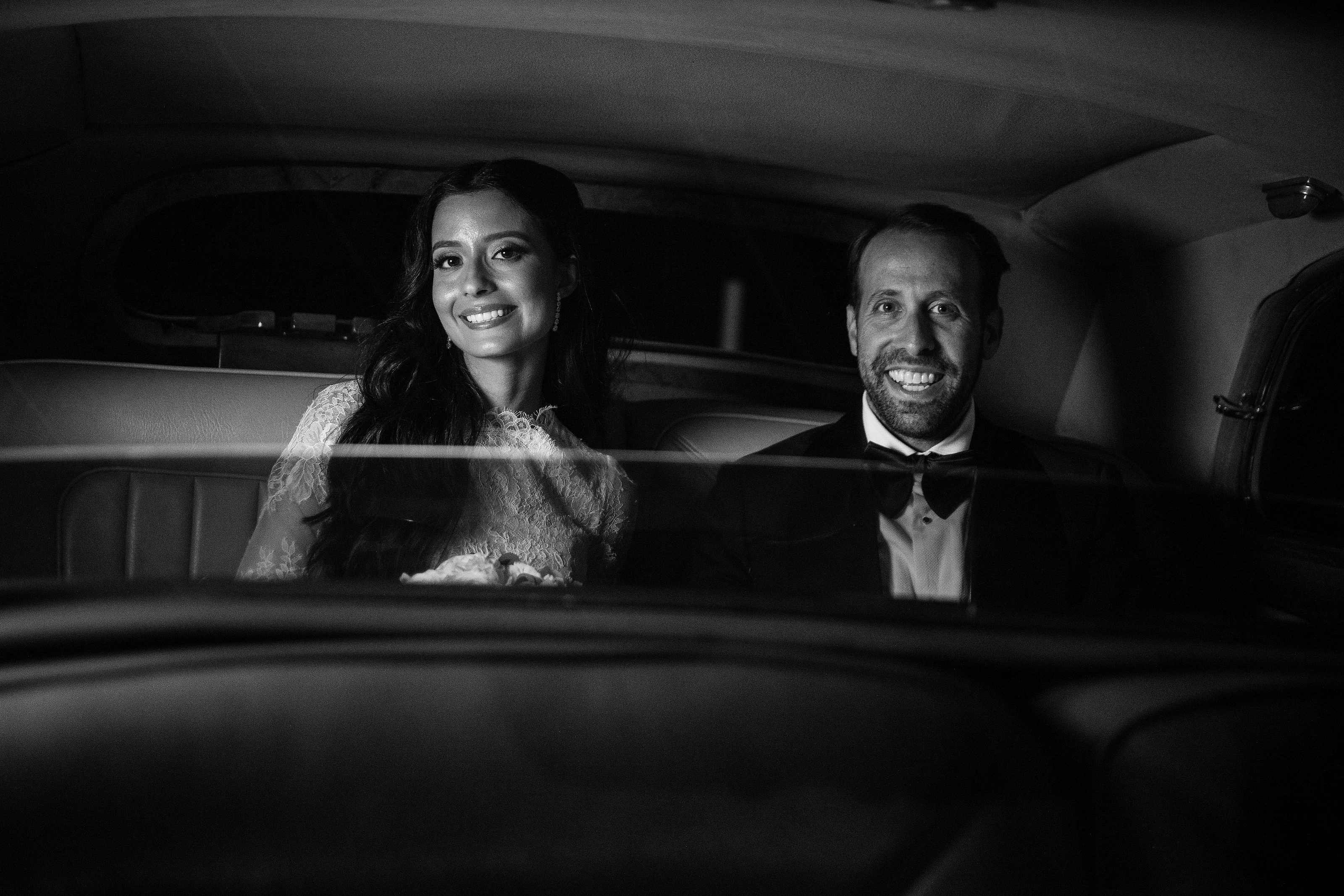 Black-and-white photo of a smiling bride and groom seated inside a vehicle, framed through the interior opening with soft overhead light.