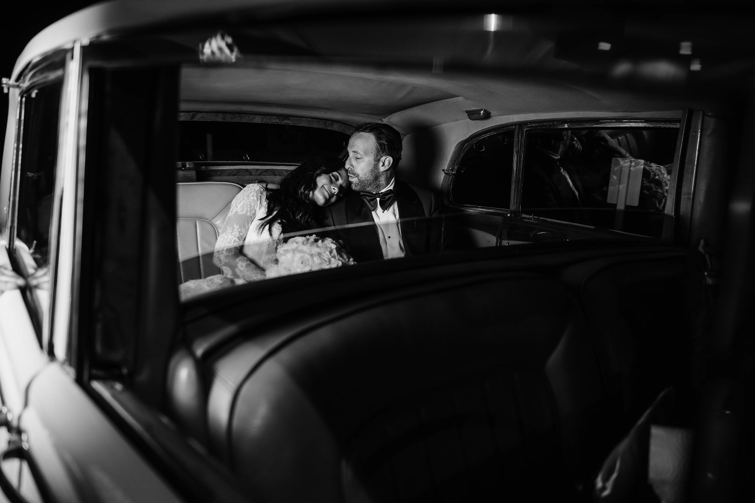 Black-and-white photo of a bride and groom sharing an intimate embrace in the backseat of a car, captured through the open door window.