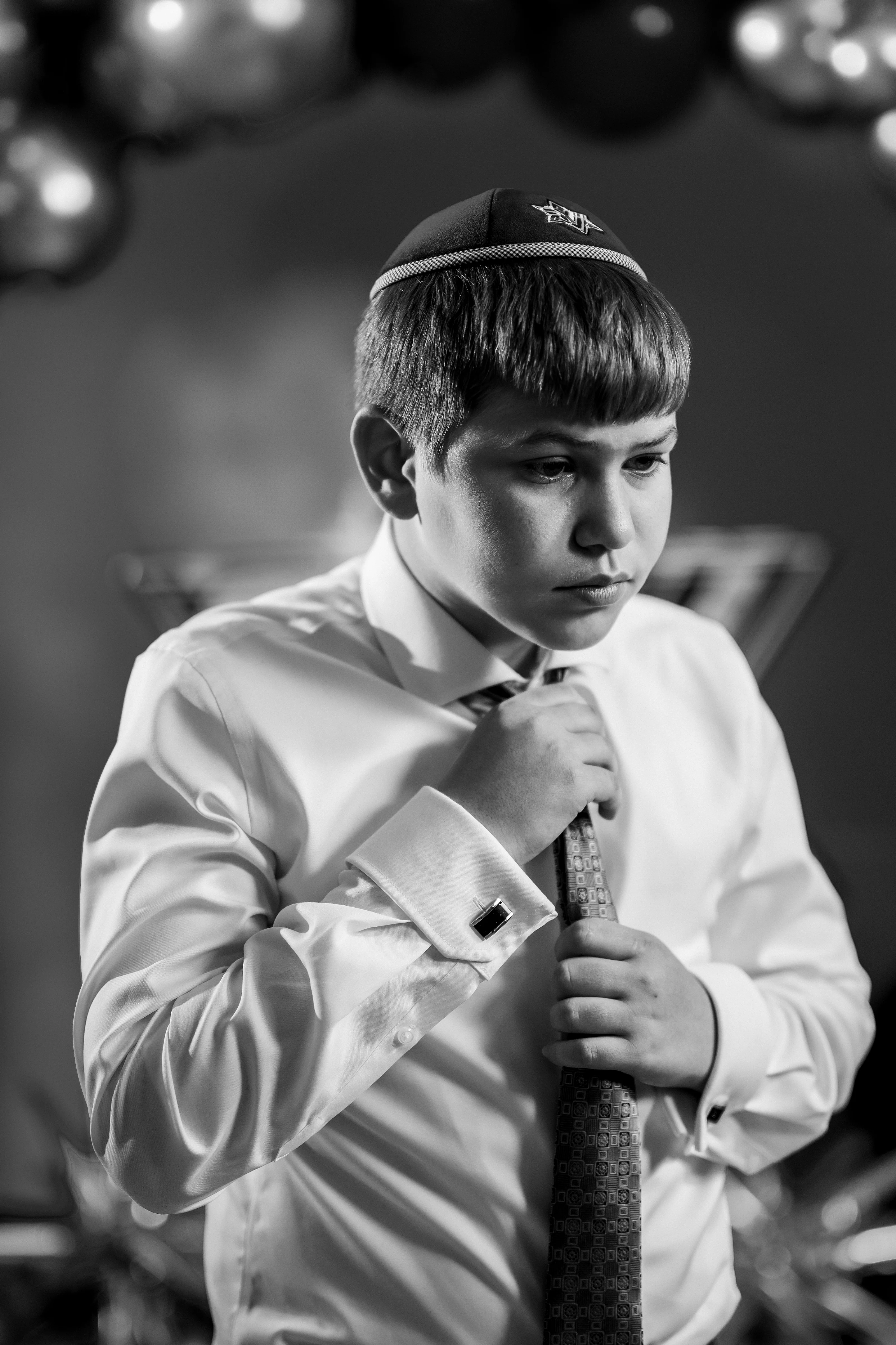 Black-and-white portrait of a young boy seated at a ceremony, wearing a kippah and white shirt and tie, adjusting his attire with a focused, solemn expression.