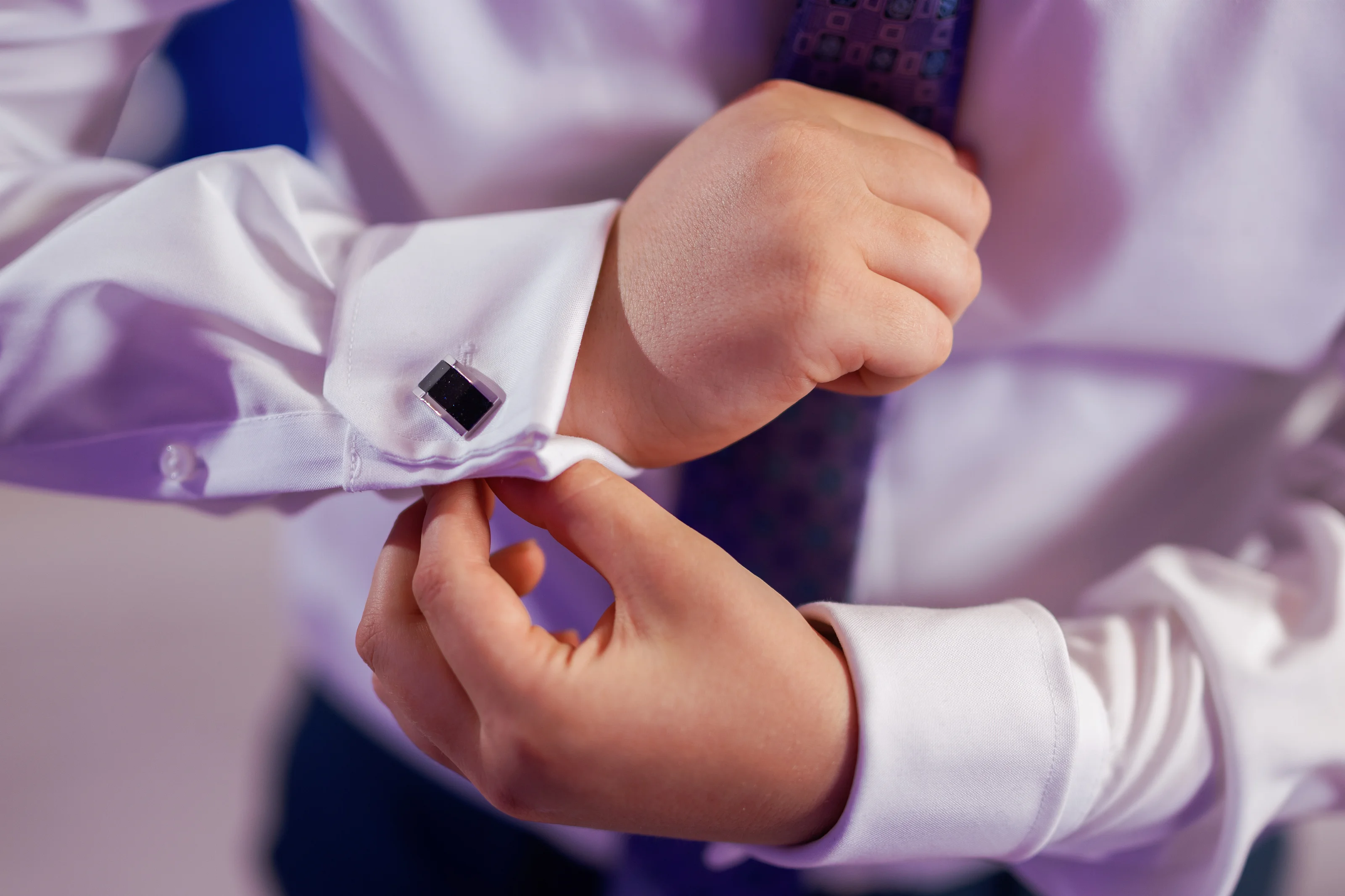 Close-up of hands placing a tefillin box and strap during a Jewish ritual, with white garments and soft pastel light in the background.