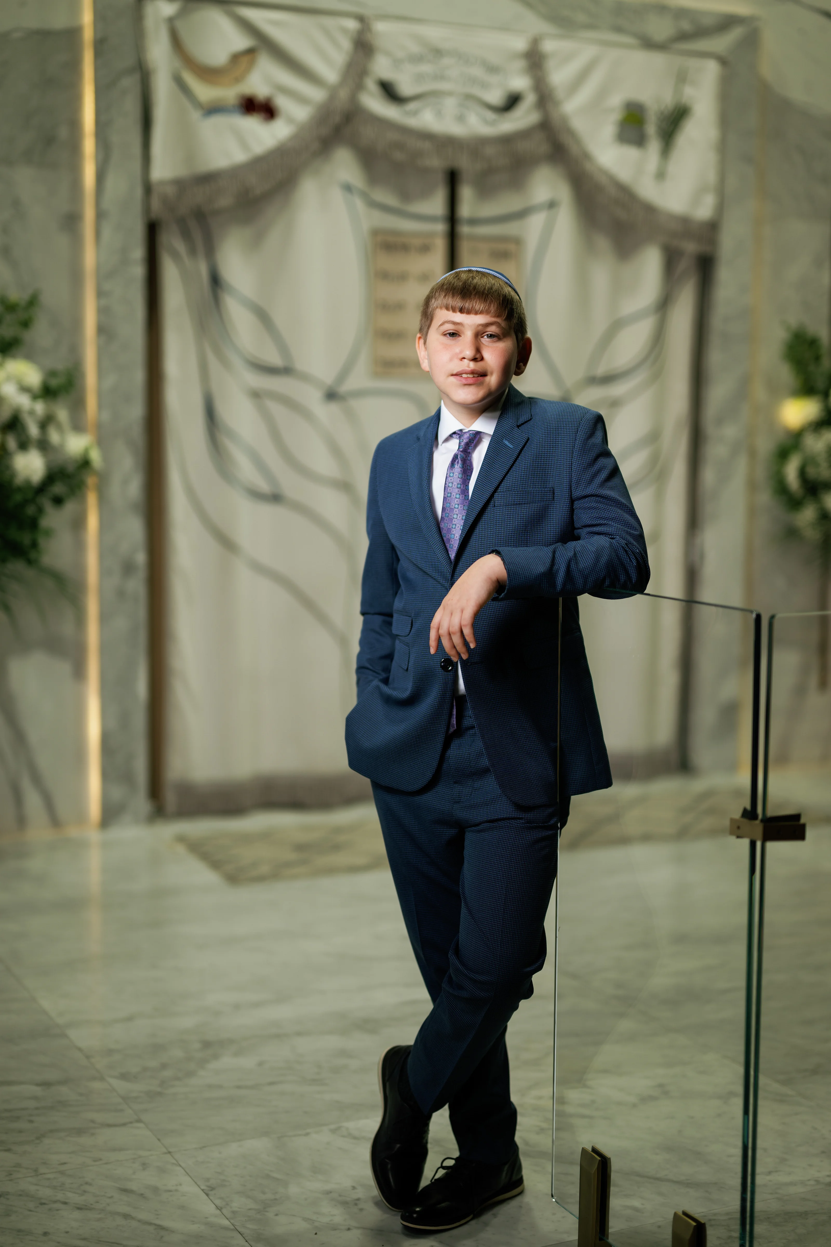 Formal portrait of a child in a suit standing with arms crossed indoors in front of an event backdrop.