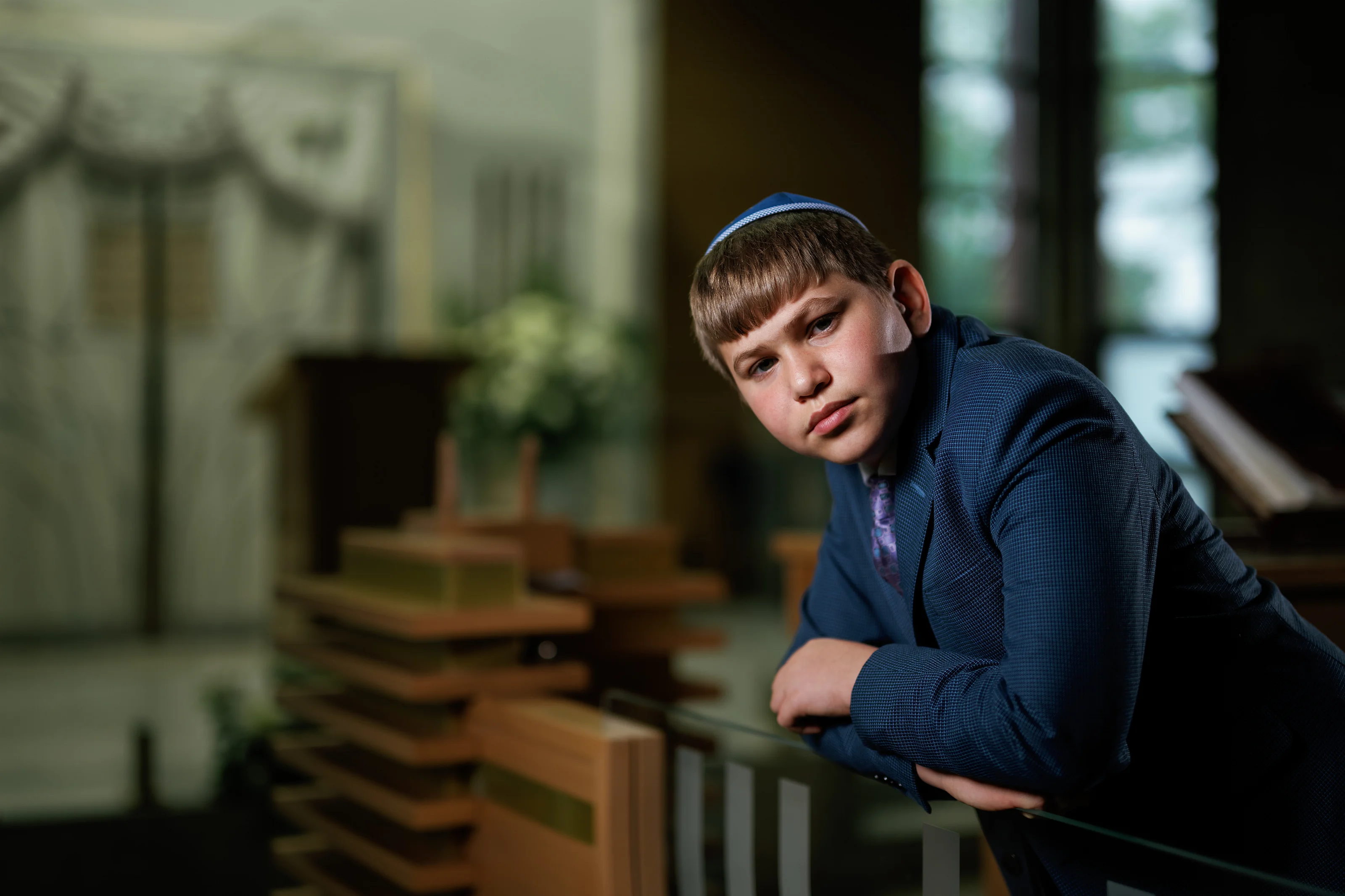 Framed indoor portrait of a young boy in a blue suit and patterned kippah leaning on a railing, with soft window light and blurred background.