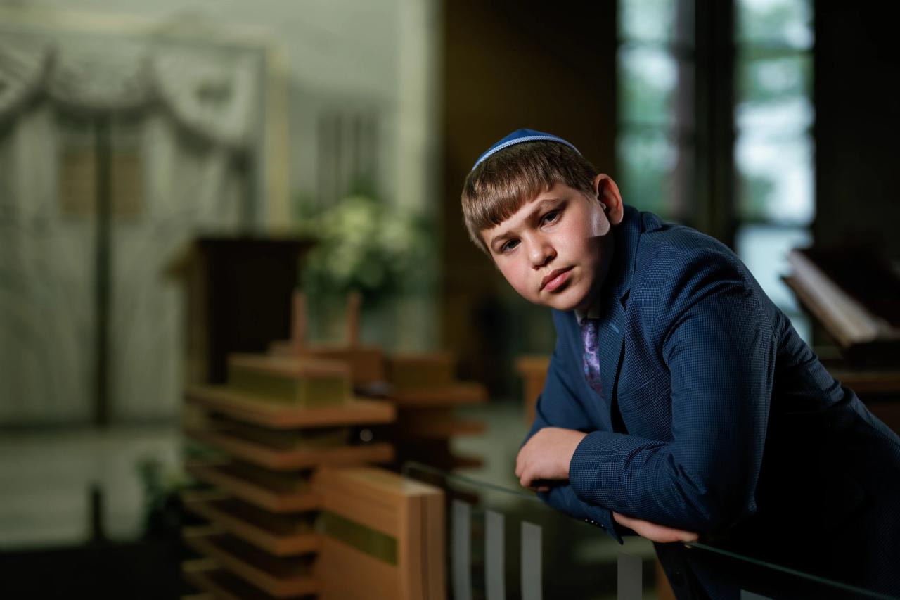 Framed indoor portrait of a young boy in a blue suit and patterned kippah leaning on a railing, with soft window light and blurred background. Bar Mitzvah photography at Congregation Ohr Torah, Valley Stream, NY.