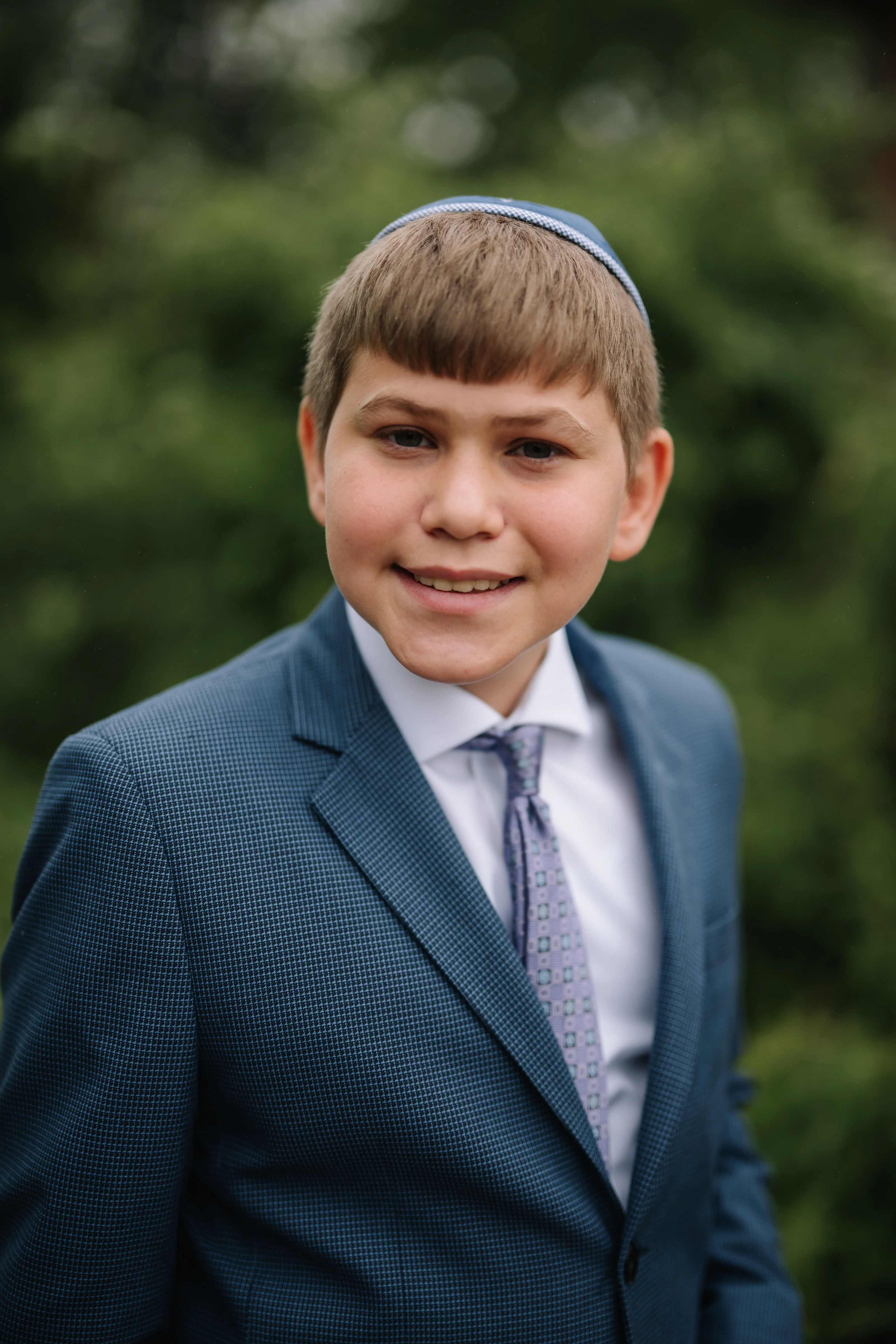 Smiling boy in a blue suit and tie wearing a light kippah-style head covering in an outdoor portrait with soft blurred greenery.