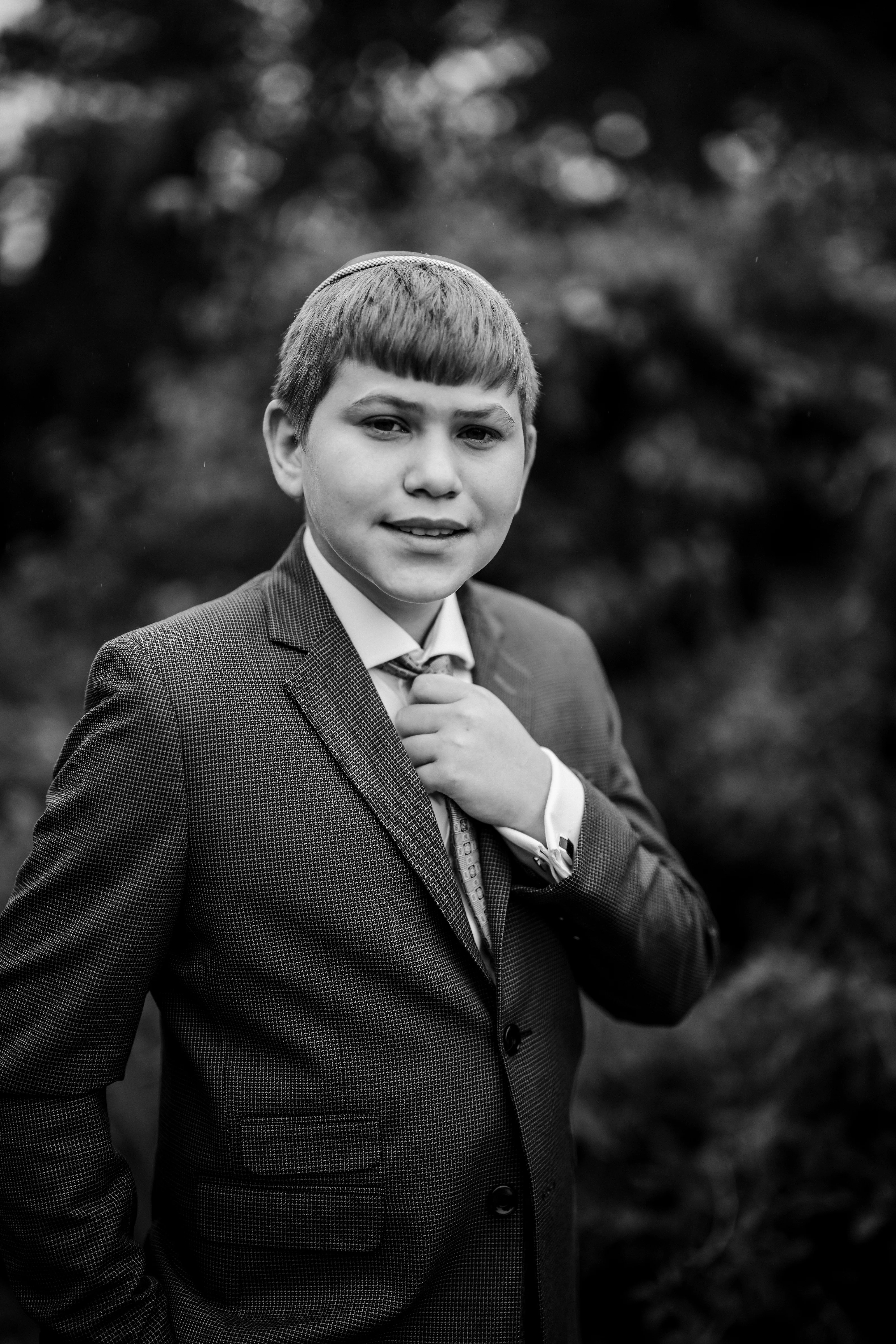 Black and white portrait of a young boy in a suit adjusting his tie, softly blurred background.