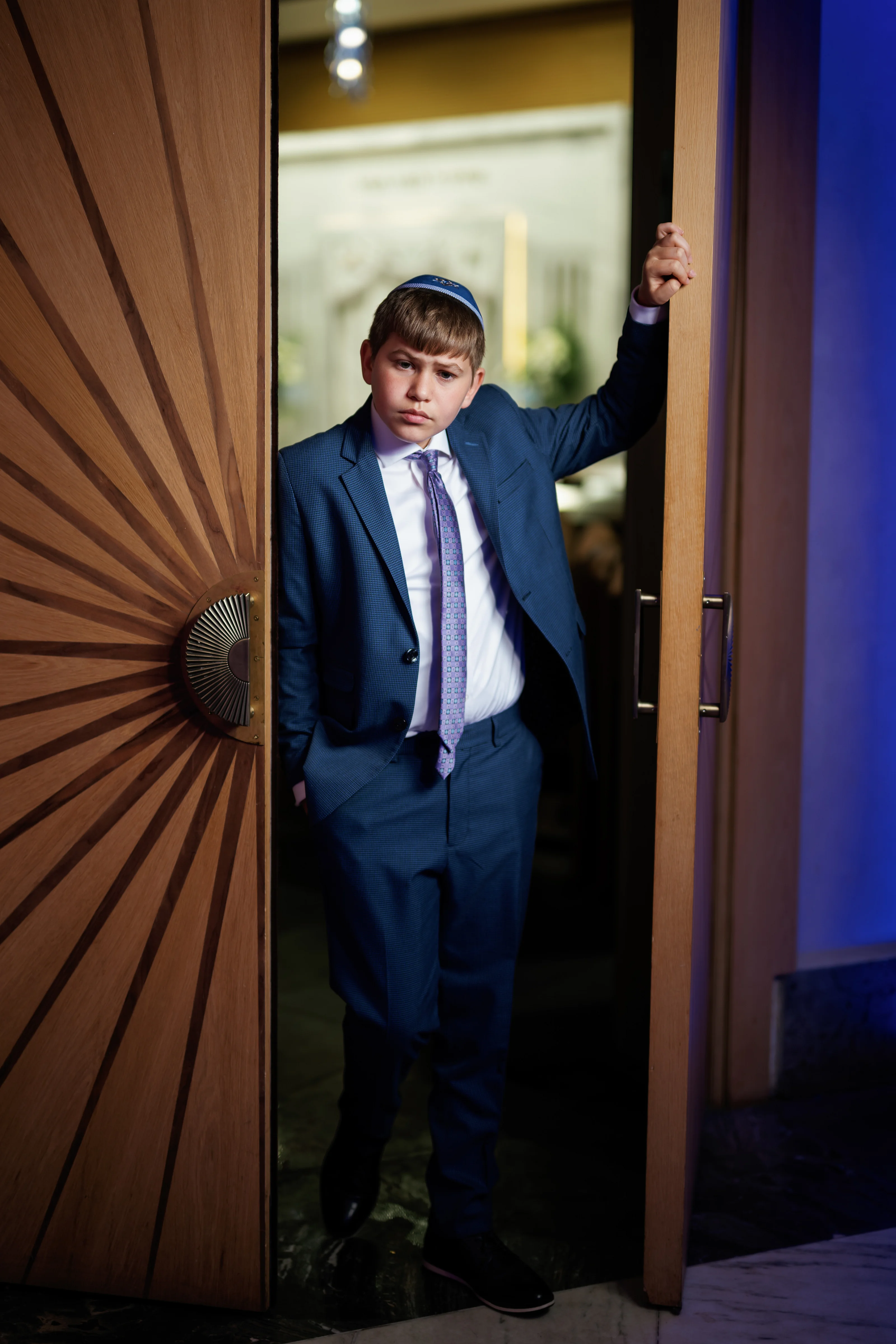 Boy in a blue suit standing in the doorway of a wooden room, one hand raised on the door, looking at the camera.