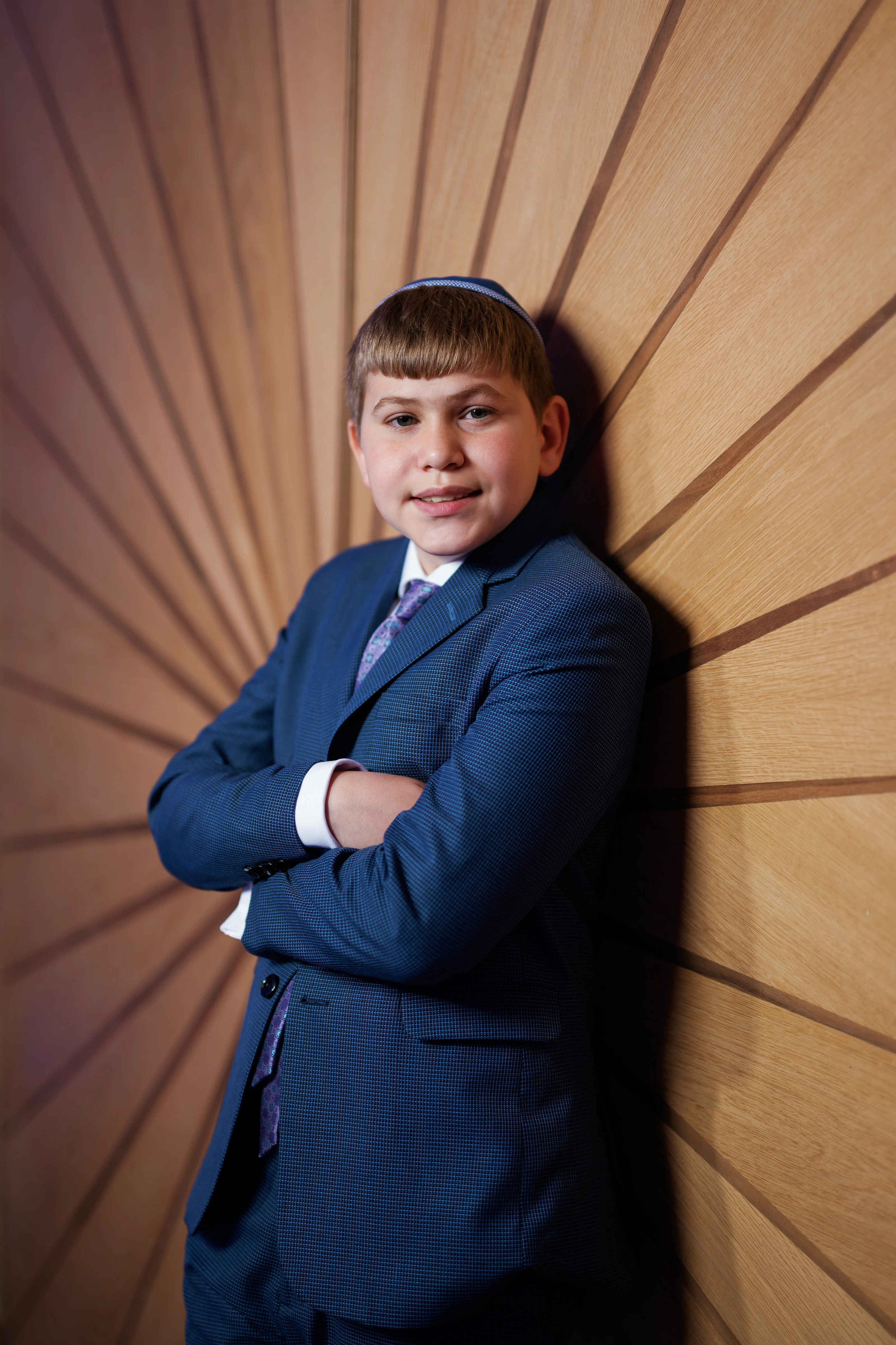 Young boy in a blue suit posing in front of a warm wooden sunburst backdrop, arms crossed and looking at the camera.